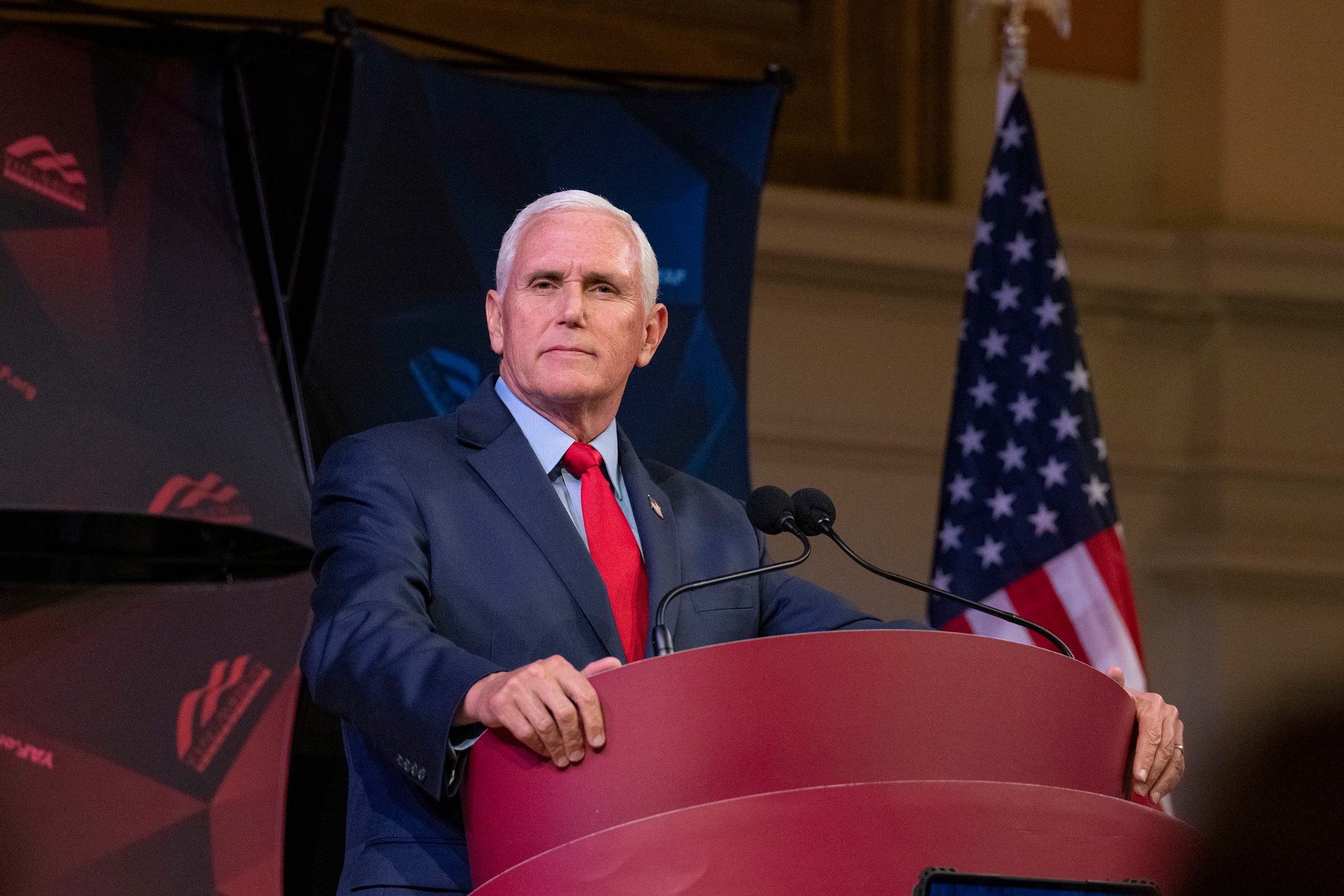 Mike Pence, wearing a blue suit and a red tie, stands at a podium, in front of an American flag.