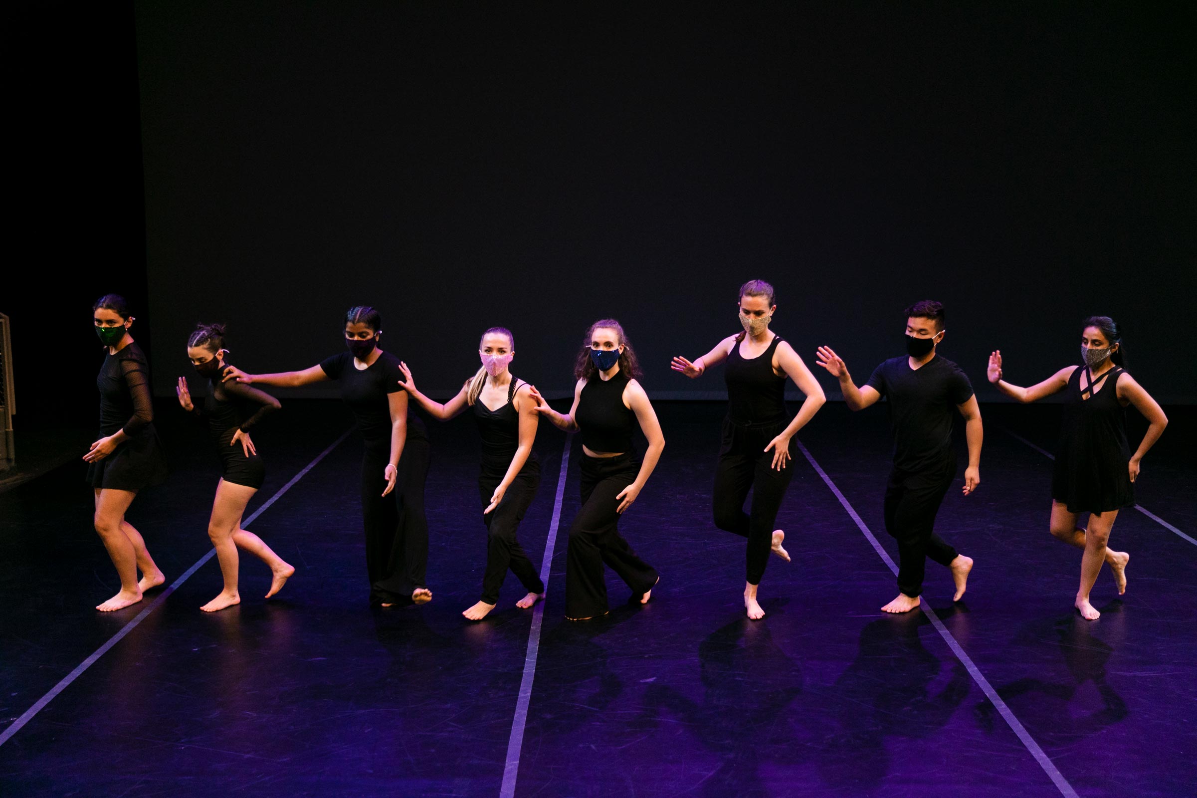 A line of dancers dressed in black, wearing masks, pose on a stage lit in purple