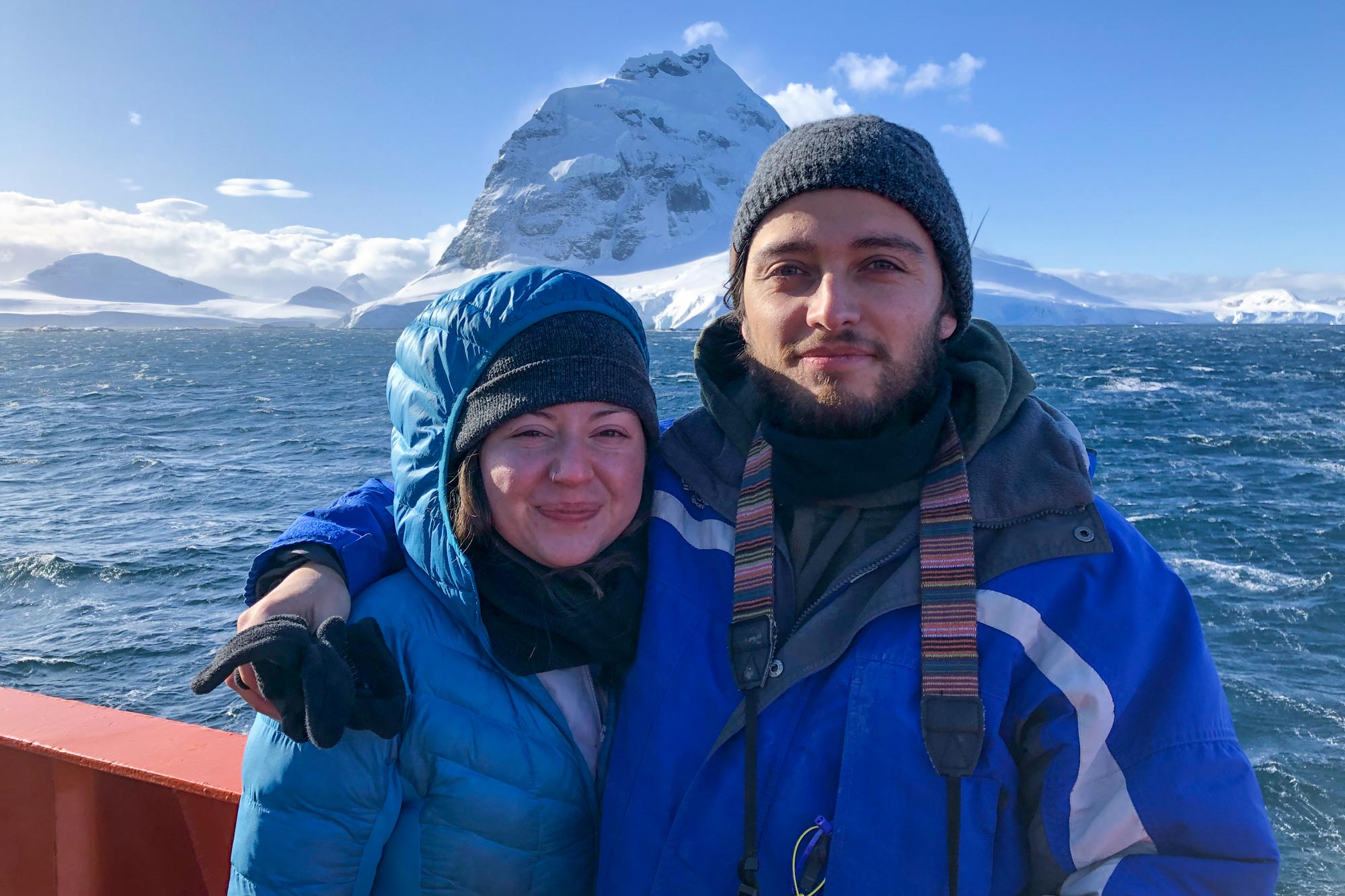 Allison Lepp and Santiago Munevar stand on a boat and pose for a photo in front of a glacial peak across the water