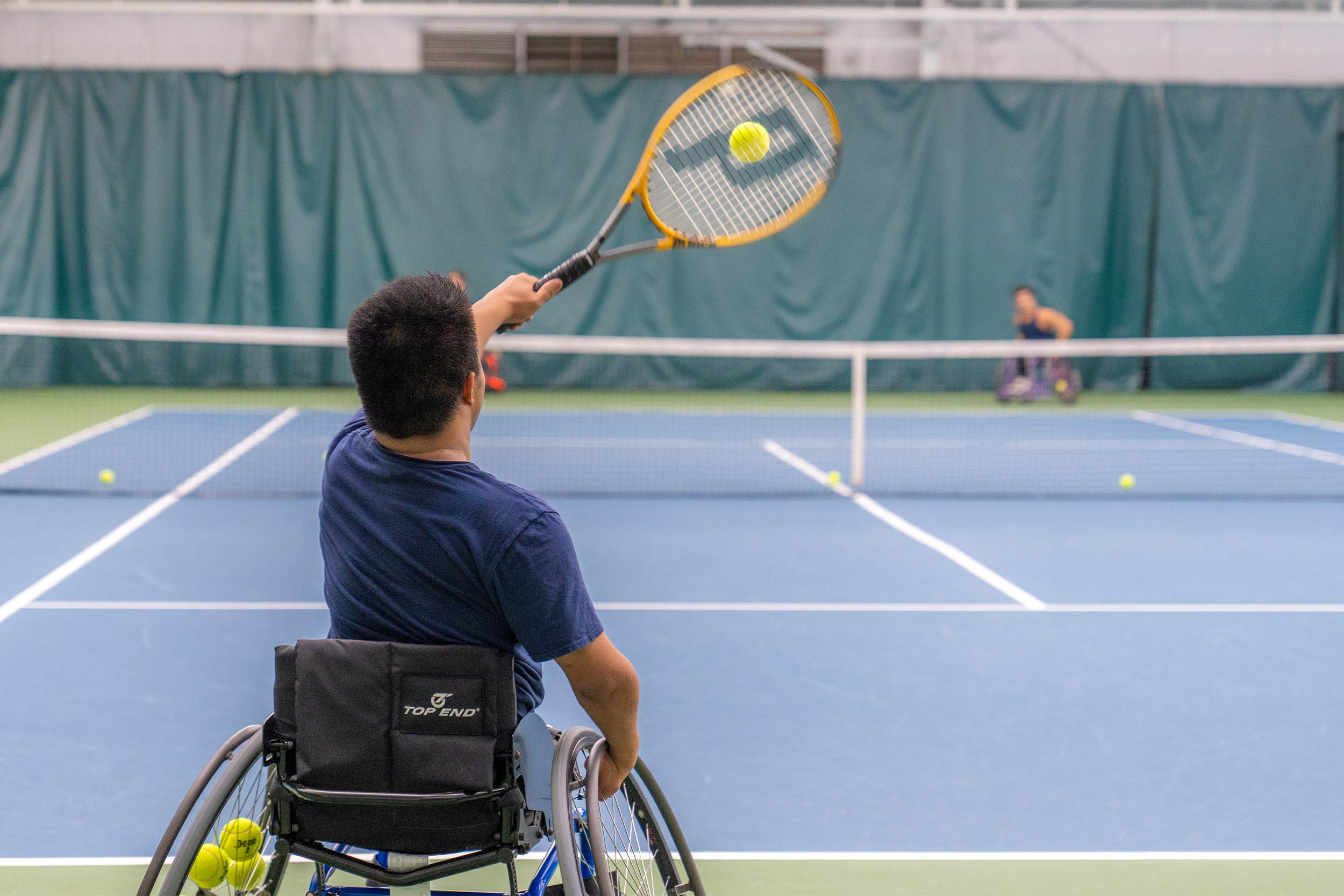 Man in wheelchair hits tennis ball during a match. In background is the other participant in the match. 