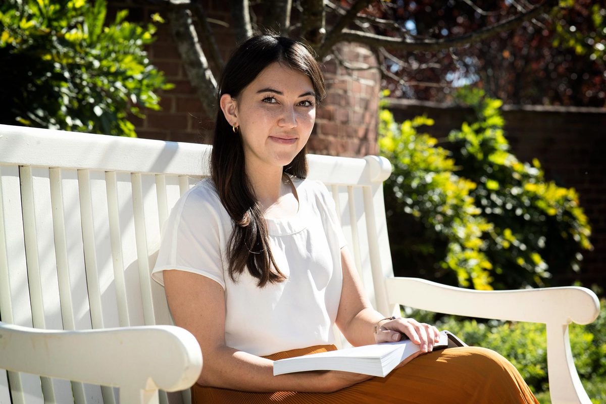 Alexandra Pentel sitting on white bench holding a book looking at the camera