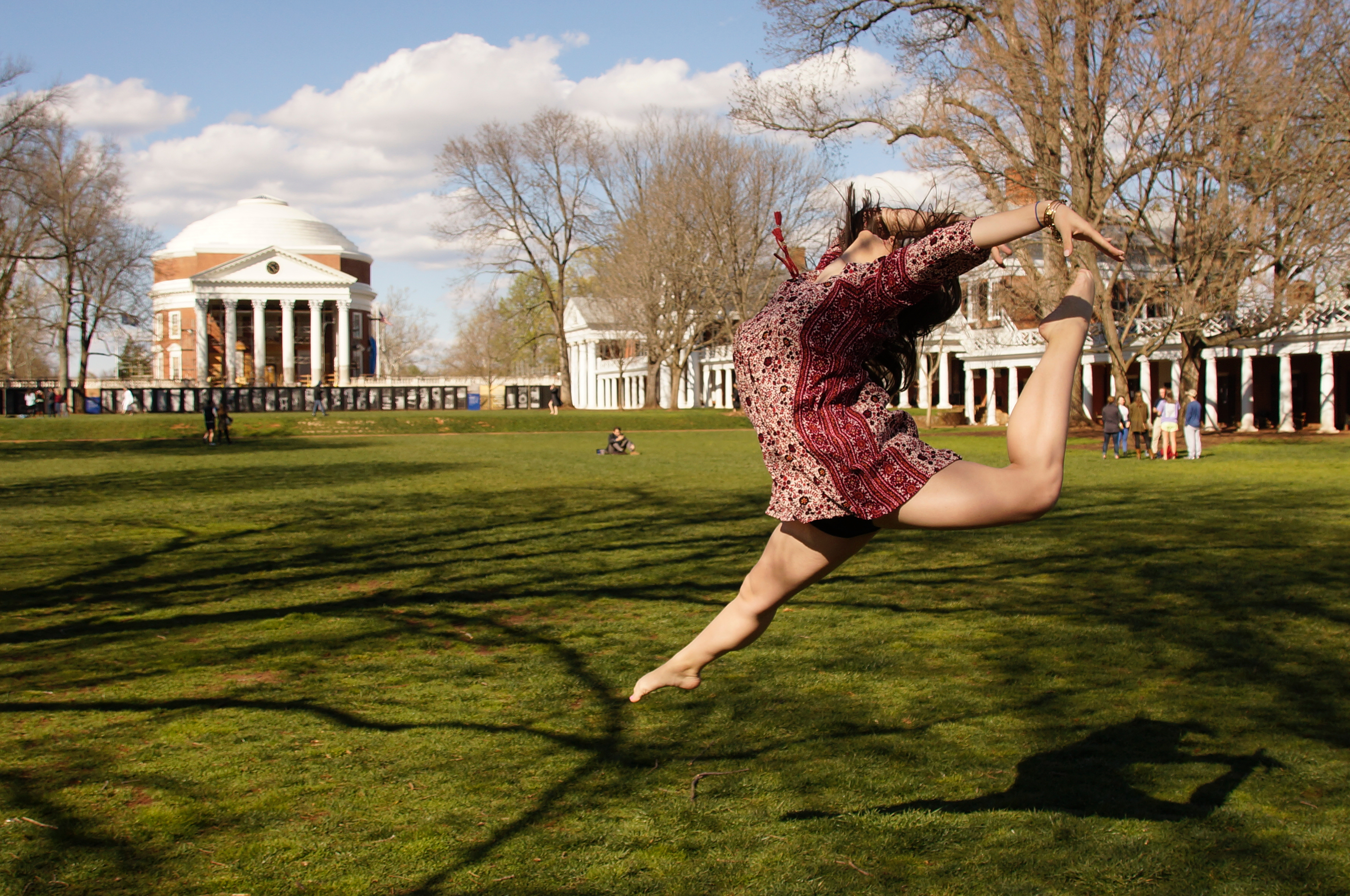 Woman on the Lawn jumps in the air tilts her head to the back and her arms out and bends one leg to almost touch her head.