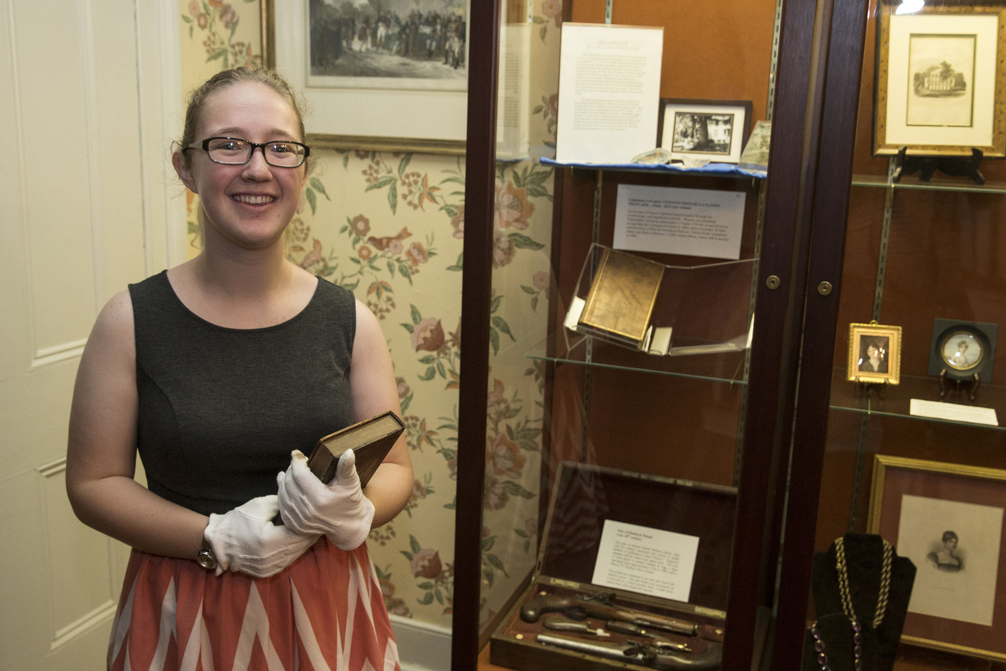 Ashley Spinks holds a book from James Monroe's house next to a cabinet while smiling at the camera