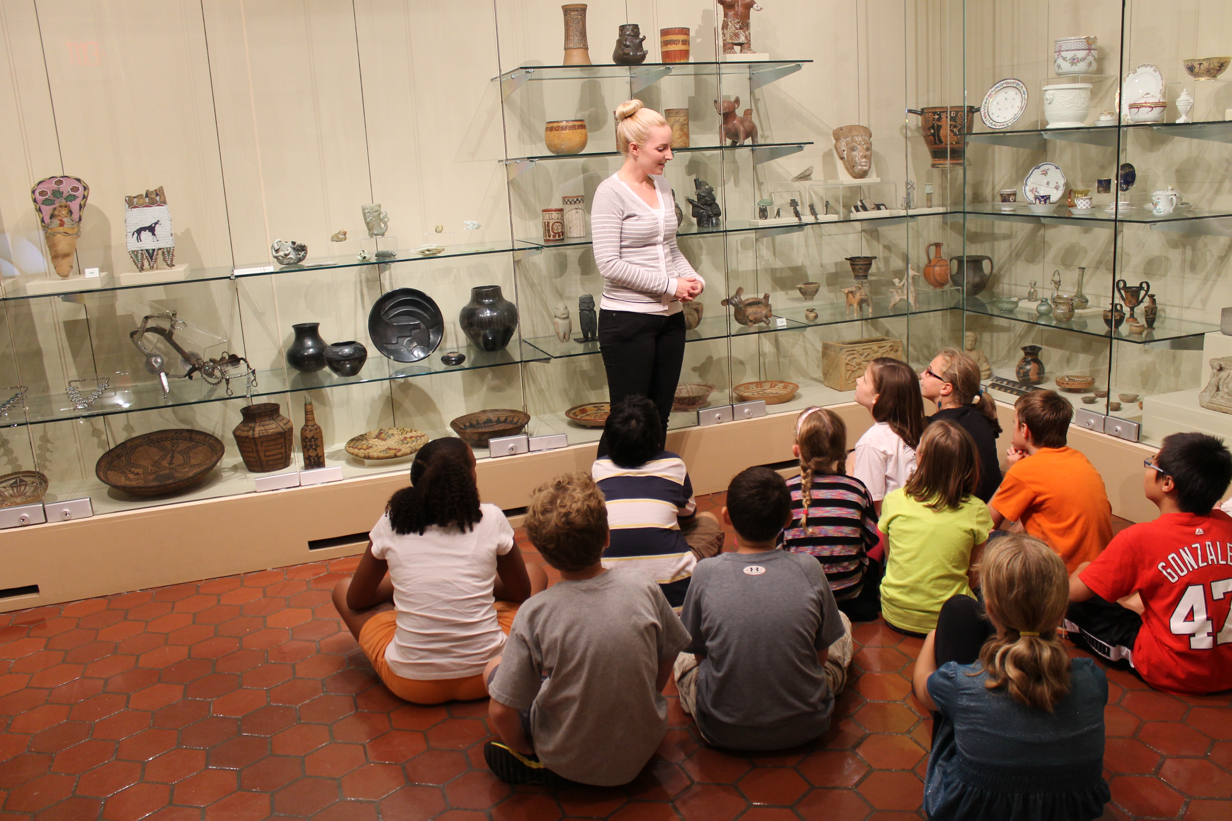 Woman stands talking about various artifacts while children sit on the floor listening