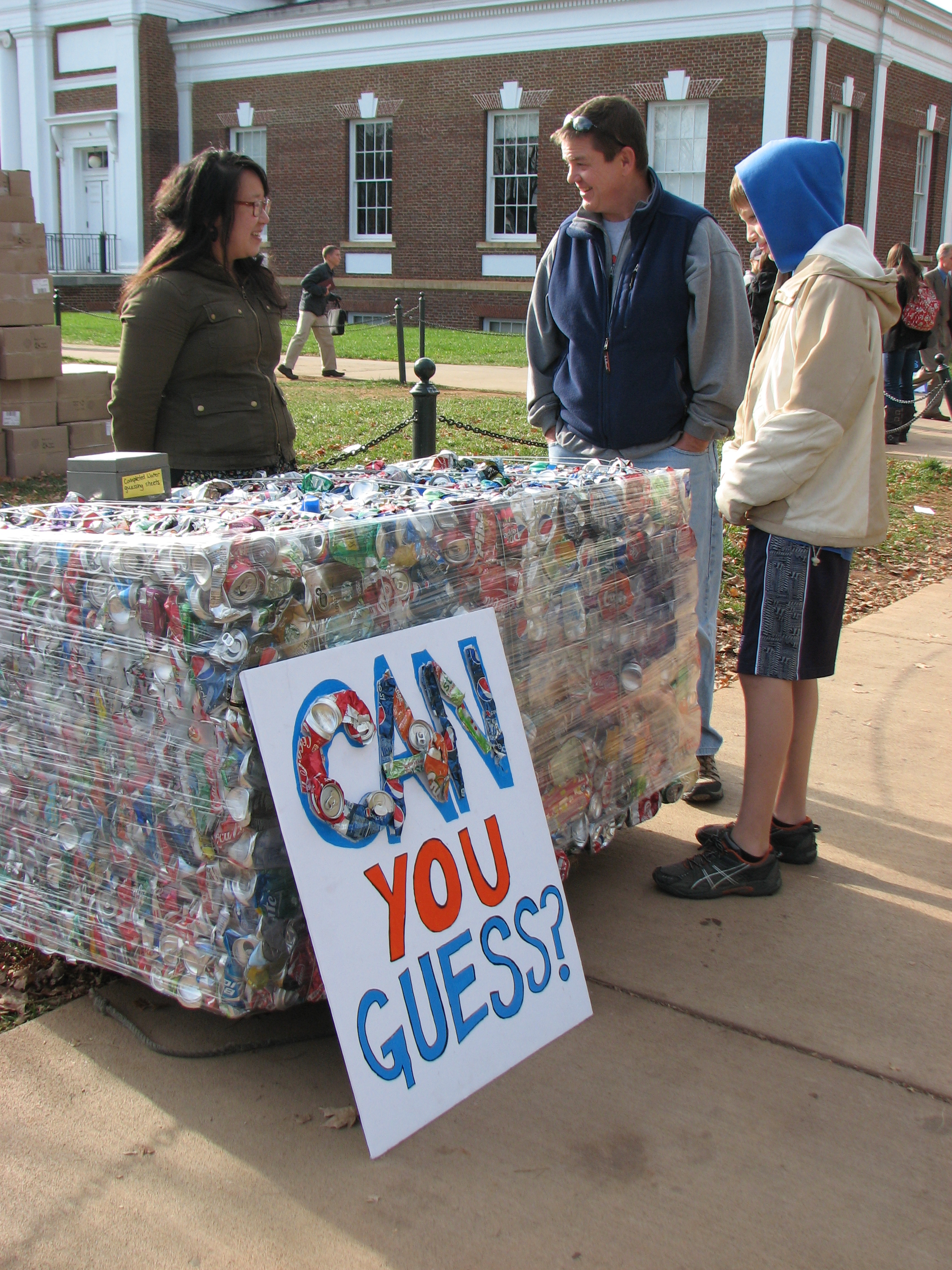 Two students talk to another student in front of a block of crushed aluminum cans