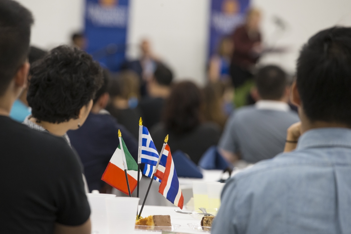 Three flags sitting on a table while people listen to a speaker at a podium at the front of the room