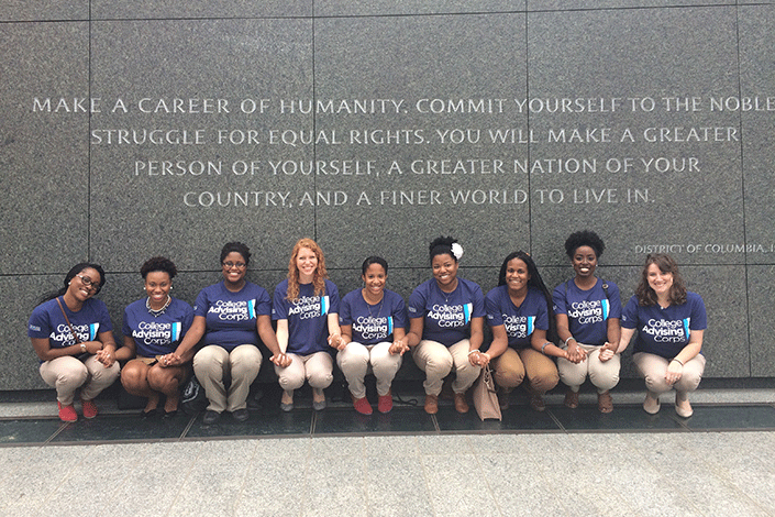 College Advising Corps participants squat under a quote engraved on a a rock wall. Quote: Make a career of Humanity. Commit yourself to the noble struggle for equal rights.  You will make a greater person of yourself, a greater nation of your country, and a finer world to live in.