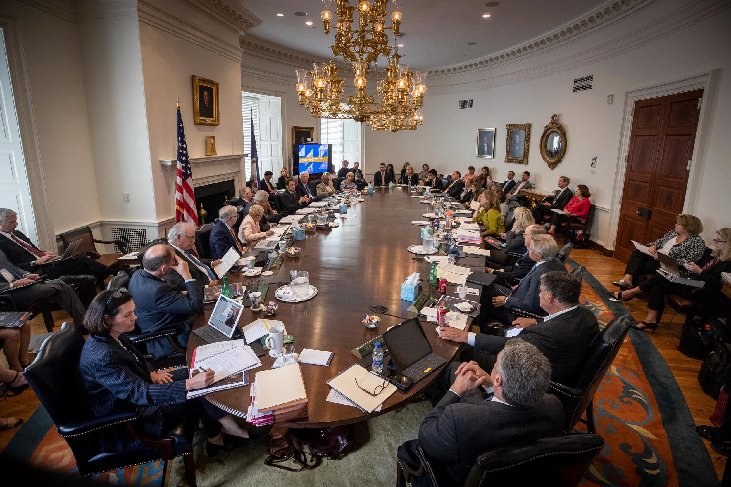 Board of Visitors sit at an oval table watching a presentation