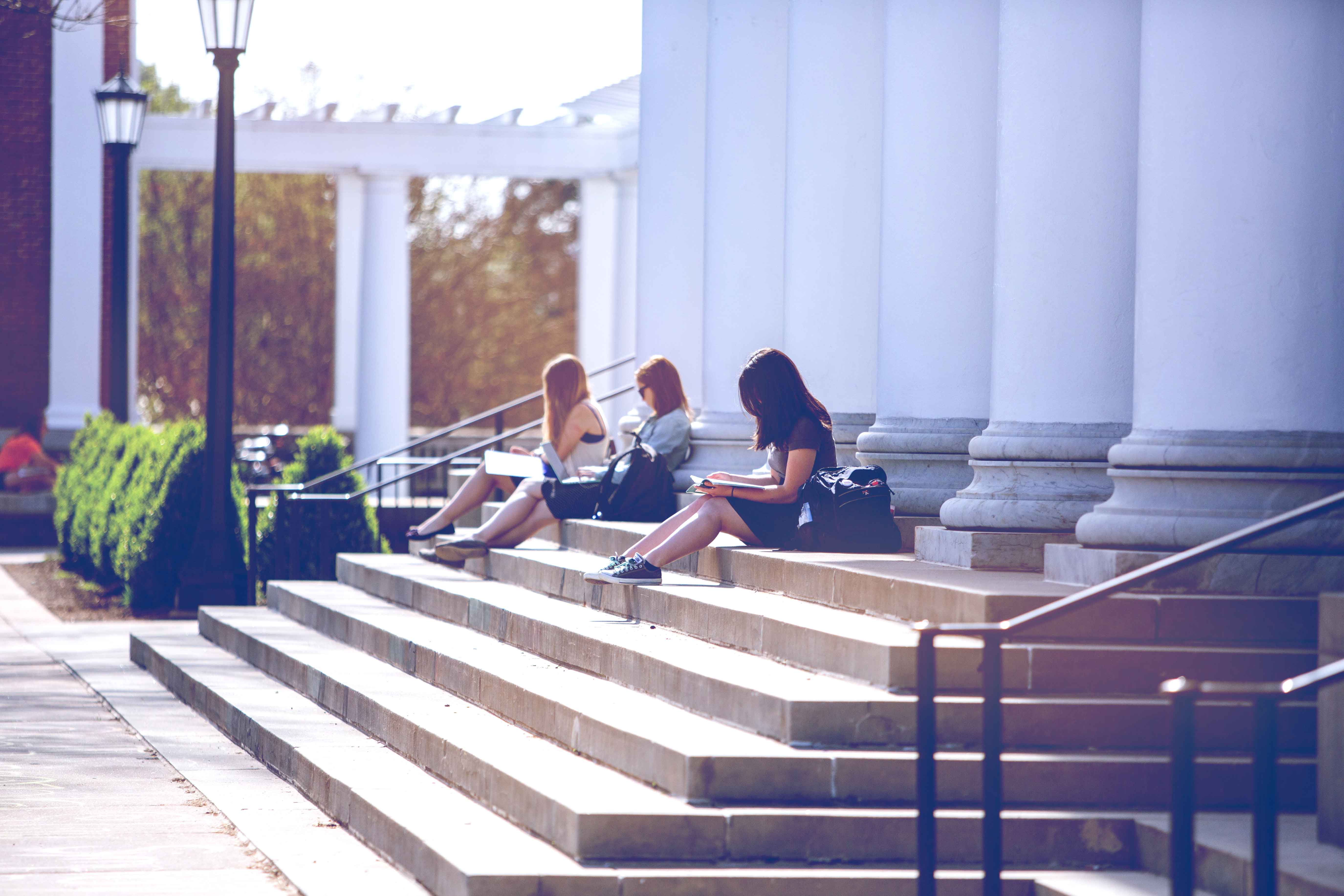 Students sitting on steps reading outside of a building on grounds