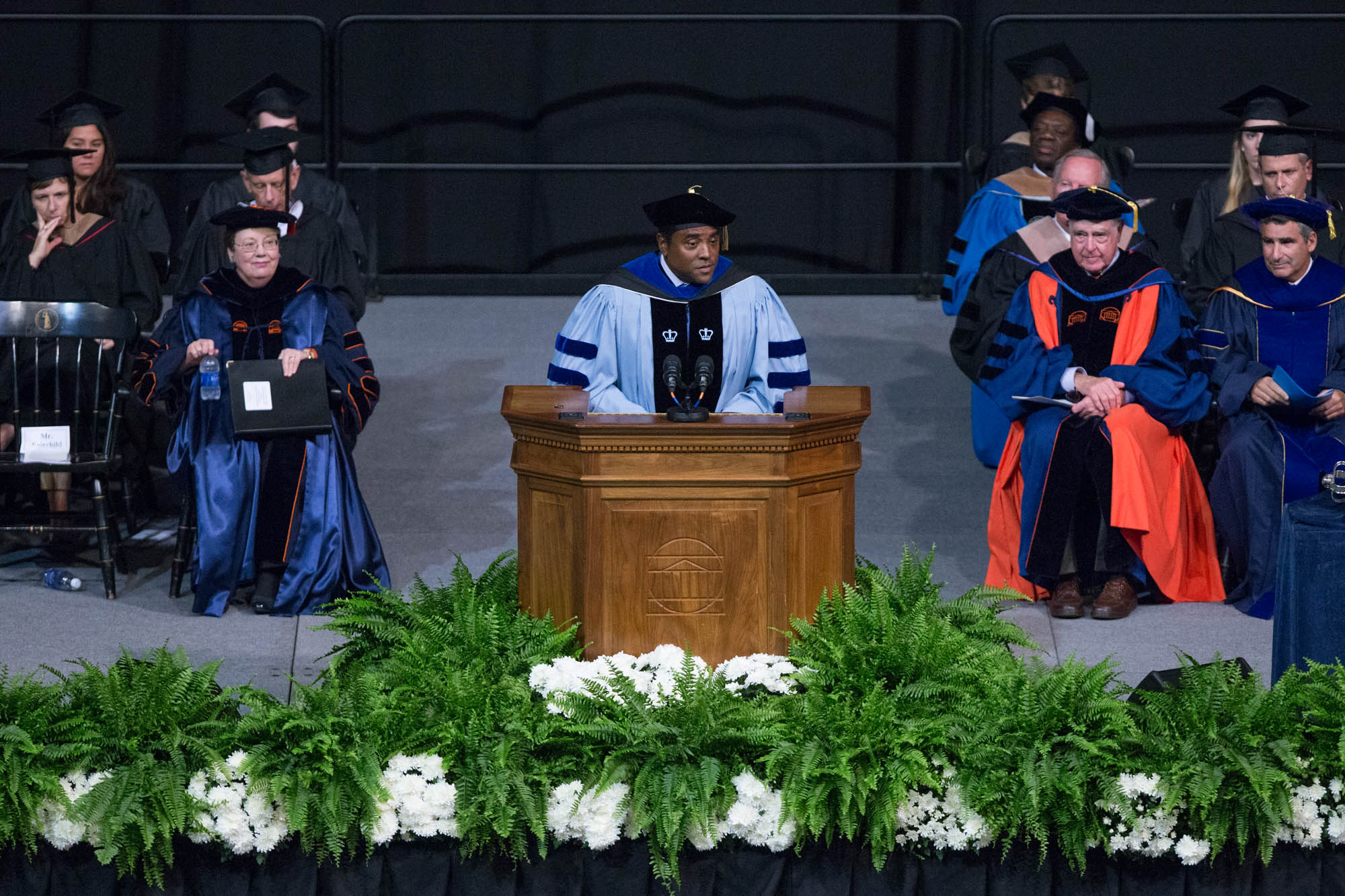 Milton Adams speaking to students from a podium dressed in graduation hat and gown