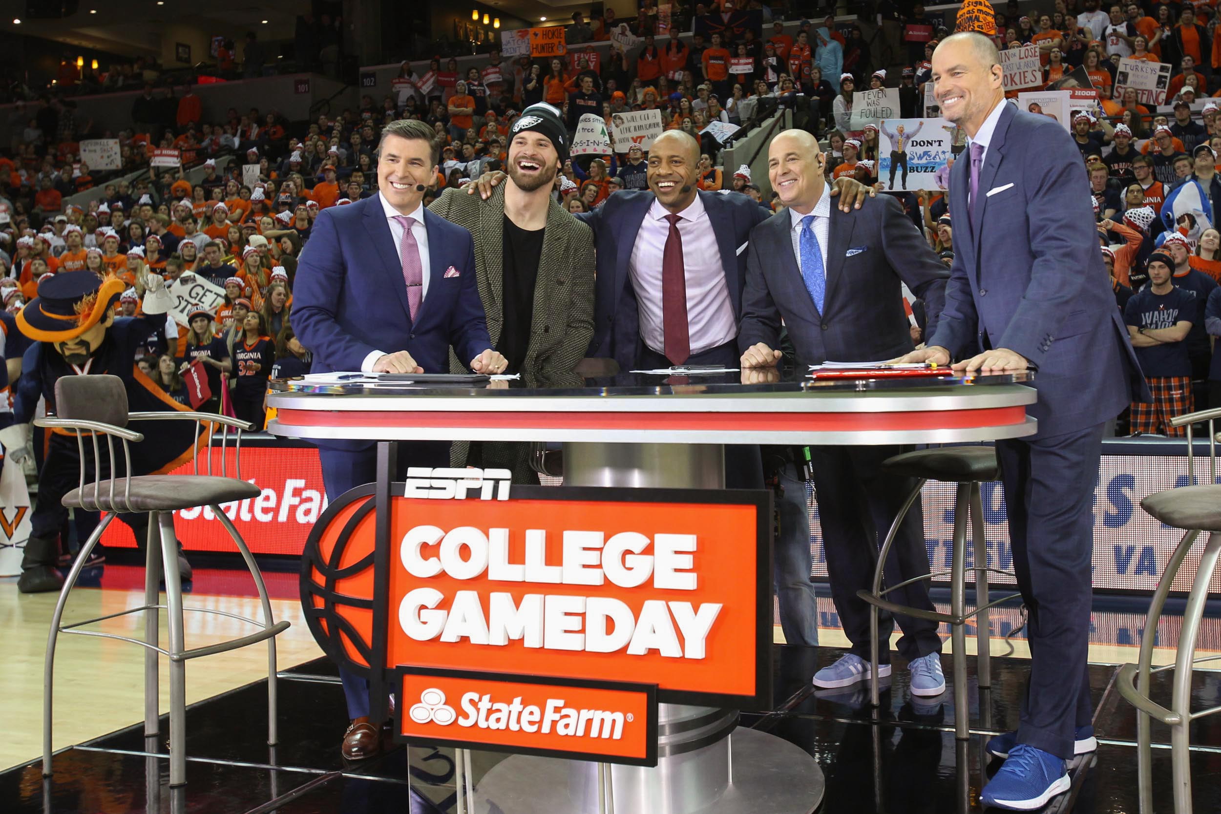 ESPN College Game Day team stands together to take a picture at their desk on the basketball court
