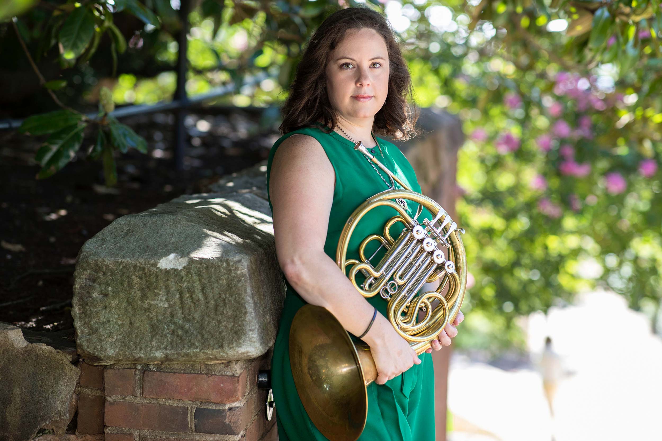 Katy Ambrose holds a french horn next to a brick wall