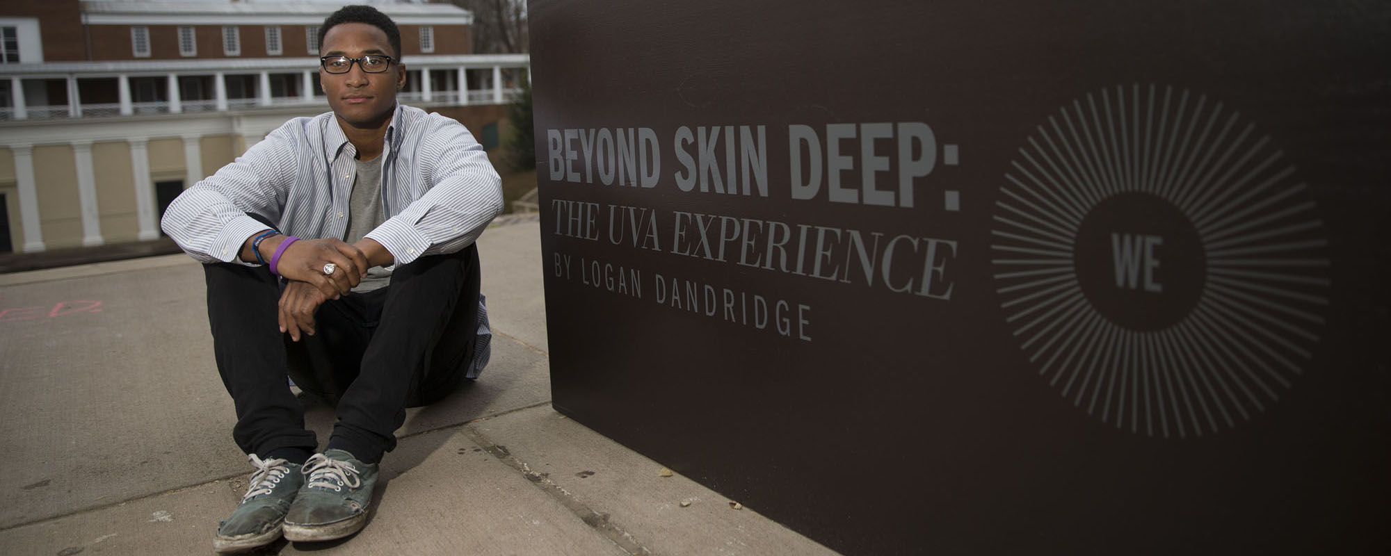Logan Dandridge sits on the ground next to a sign that says Beyond Skin Deep:  The UVA Experience By Logan Dandridge