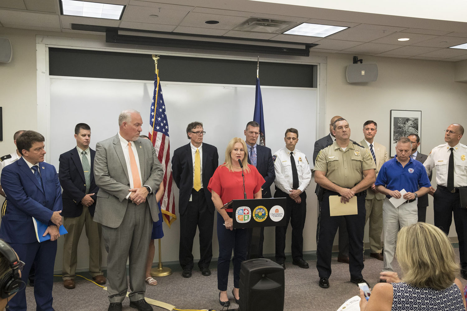 Group of UVA, state and local law enforcement officials stand at the front of a room with a woman at a podium speaking to a crowd