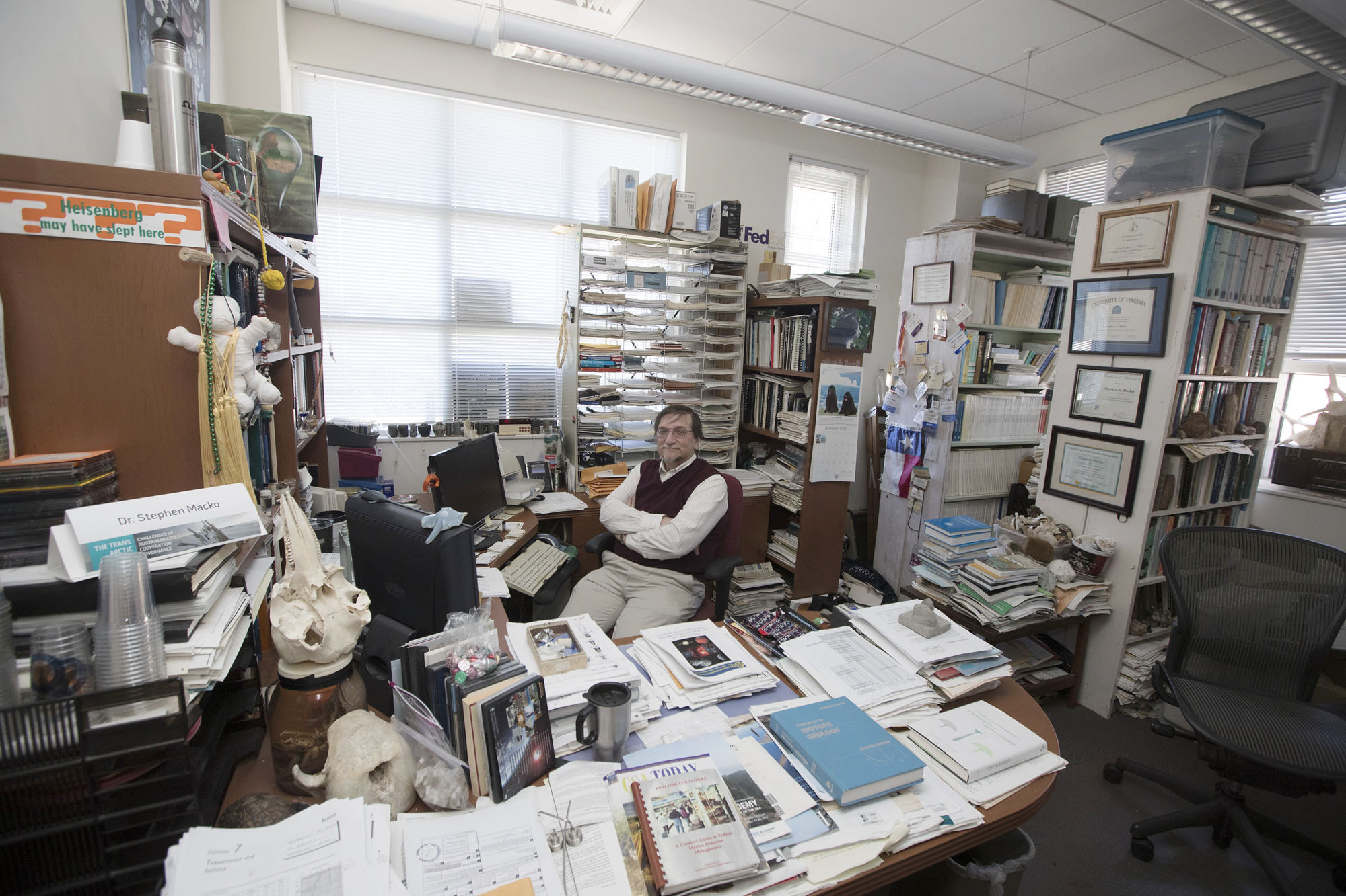 Steve Macko sits at this desk surrounded by books smiling at the camera