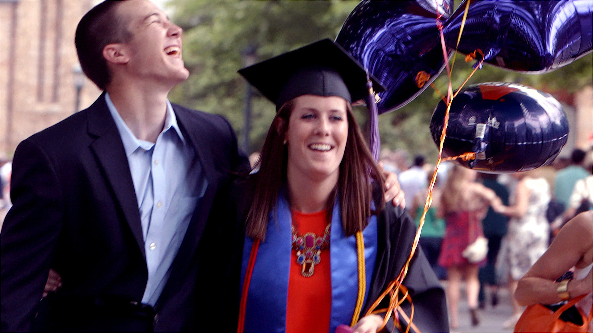 Graduate and loved one with arms wrapped around each other walking and laughing.  Graduate is carrying purple balloons