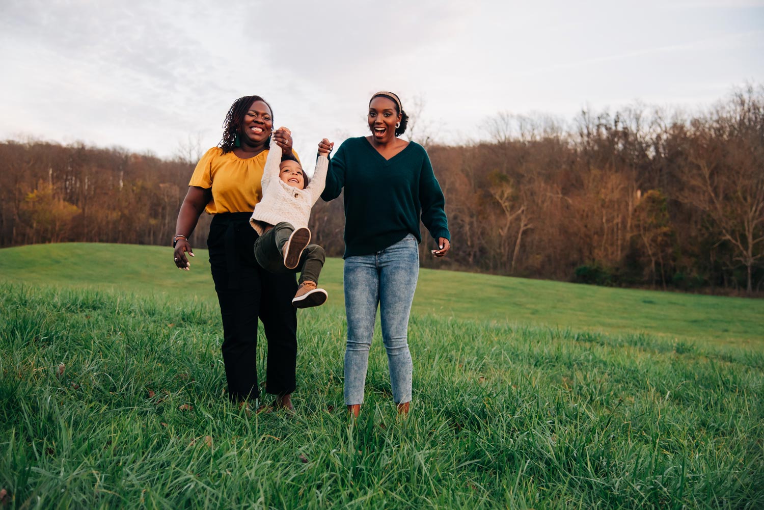 Carla Hallman, left,  and her sister, right, swing Hallman's son, center, while walking through a grassy field surrounded by trees