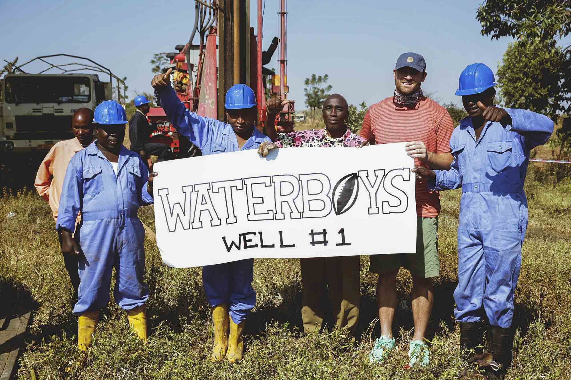 Chris Long stands with men at a well drilling site and holds a sign that reads: Waterboys well #1