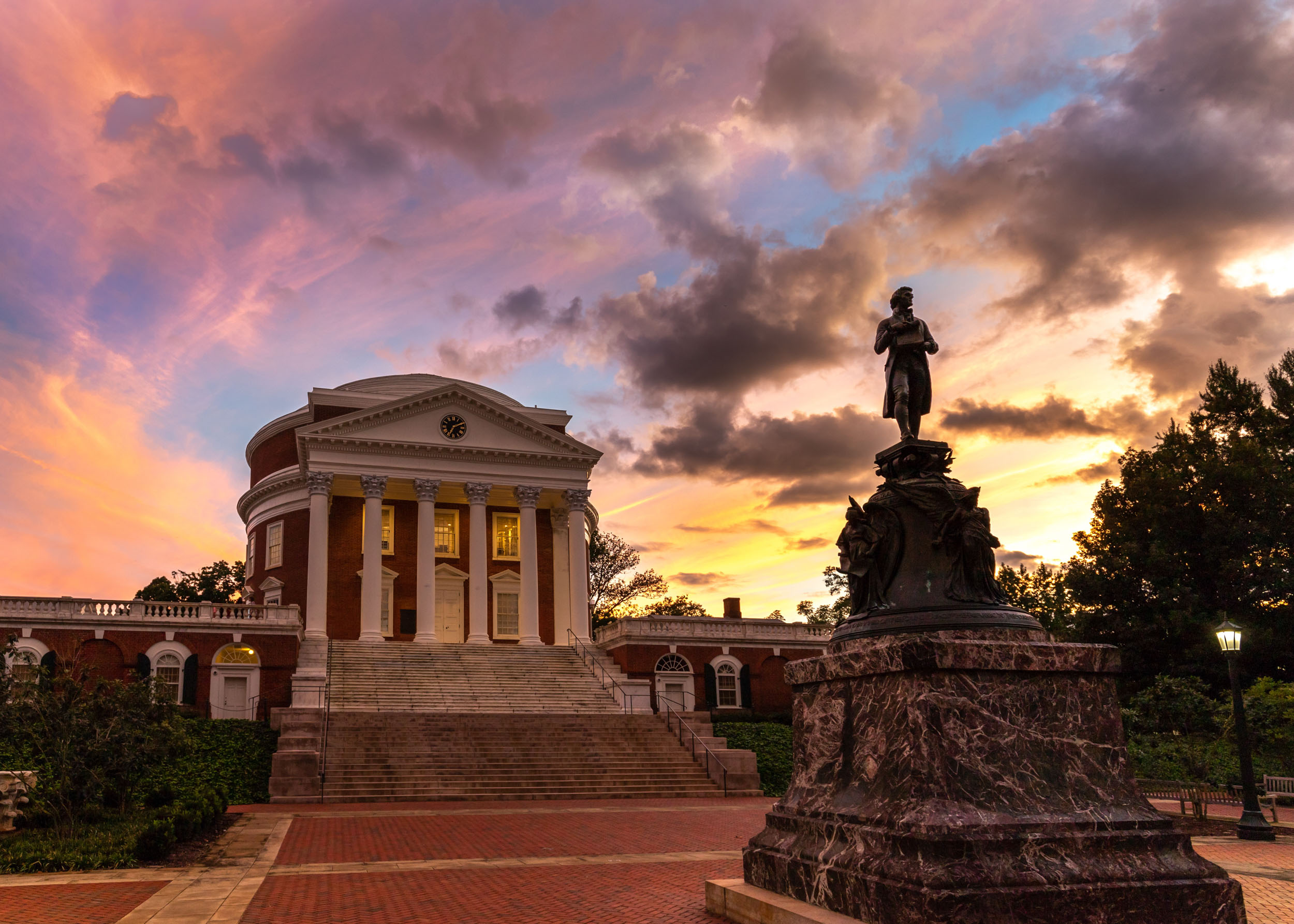 Thomas Jefferson Statue and the Rotunda are surrounded by grey clouds and a pink, yellow, purple, and blue sky
