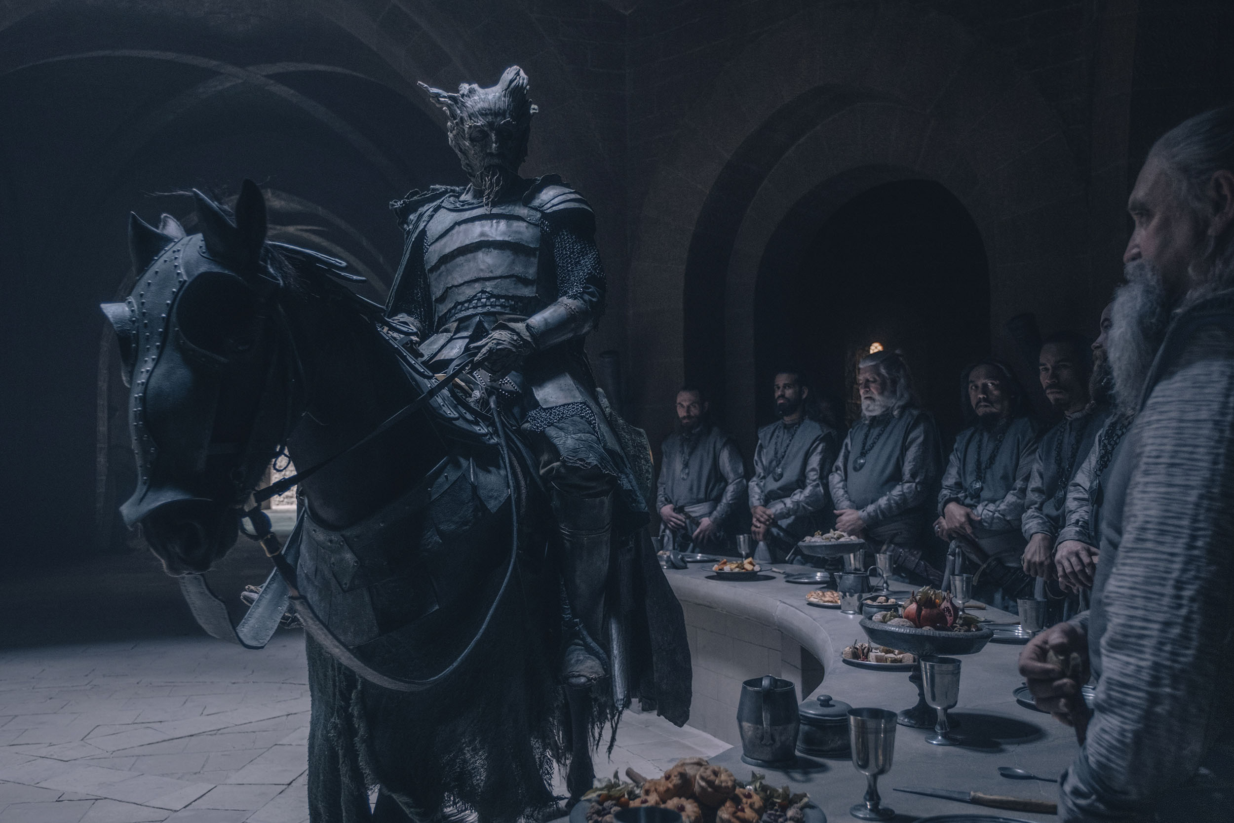 Image from the movie the Green Knight with King Arthur on his horse riding in front of the knights at an arced table