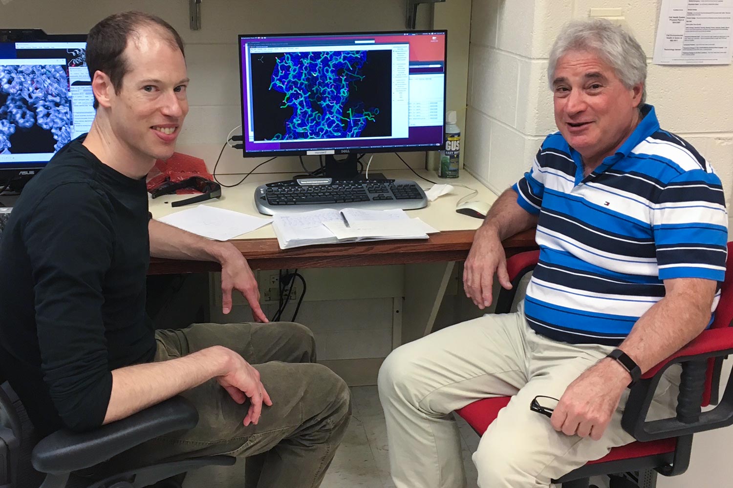  Dr. Peter M. Kasson and Edward H. Egelman sit in chairs looking at the camera with a blue structure of a virus behind them