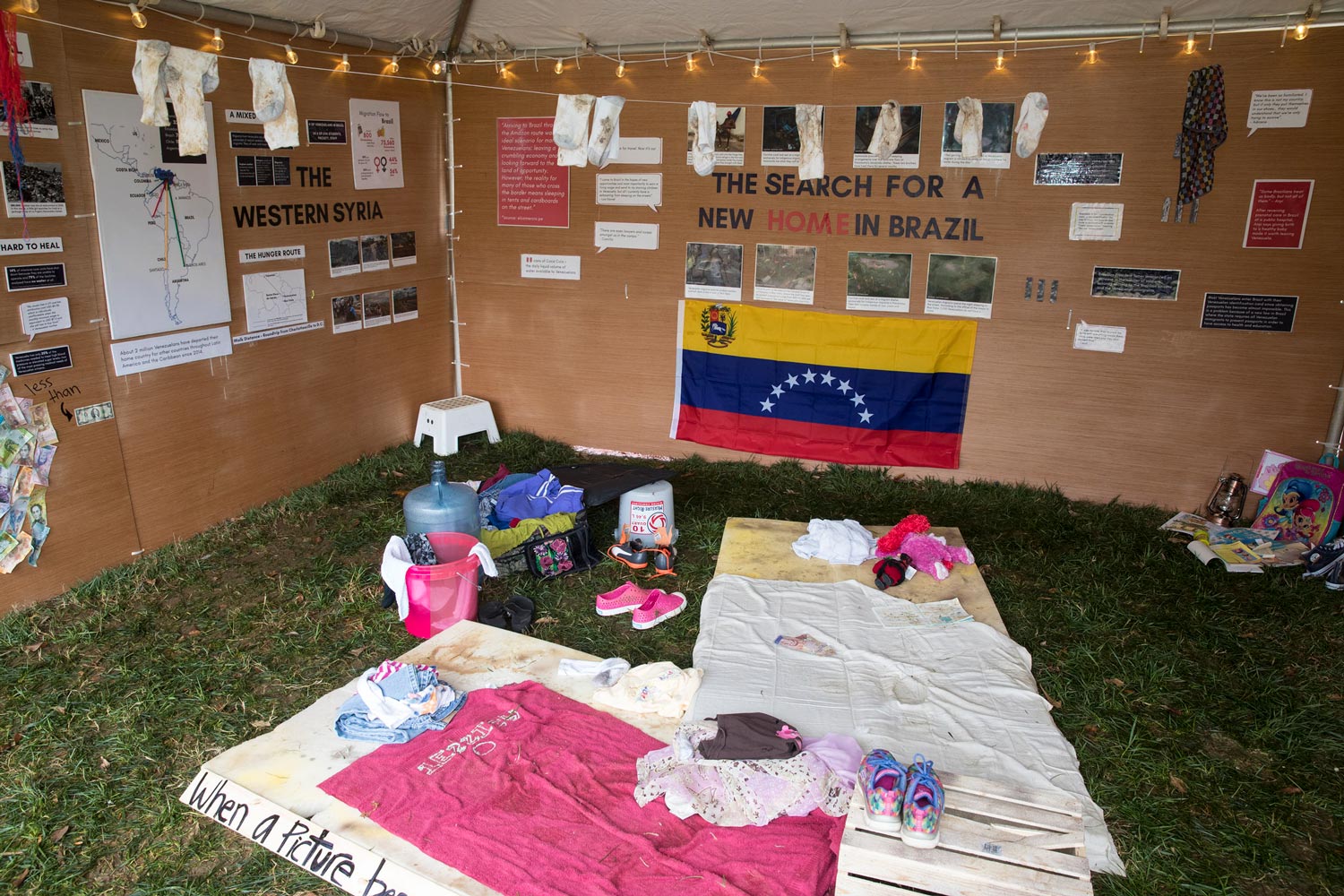Tent with cardboard walls that has text, images, and dirty socks handing up.  On the ground is personal items and sleeping mats