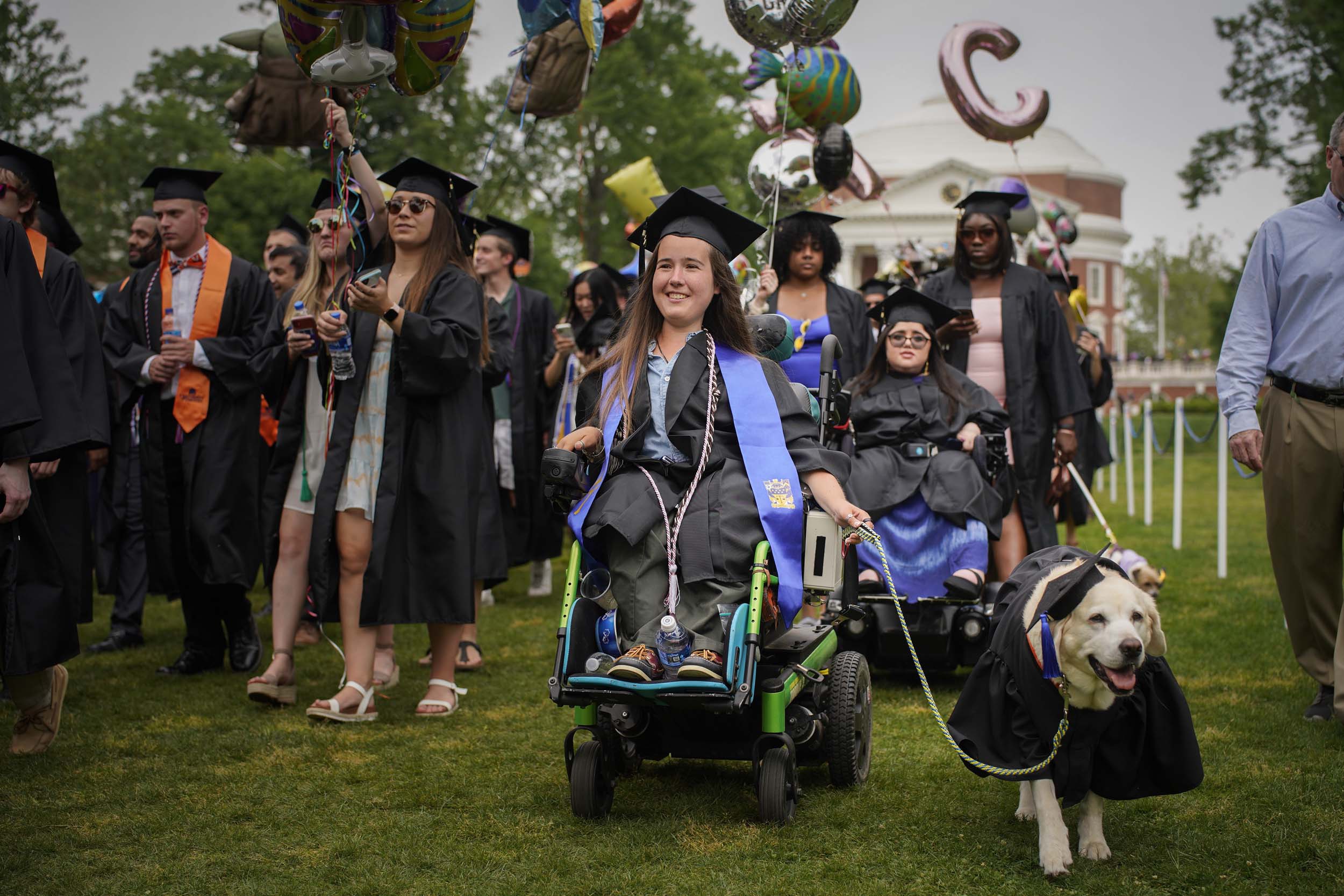 Graduates walking in and a Service Dog dressed in a cap and grown