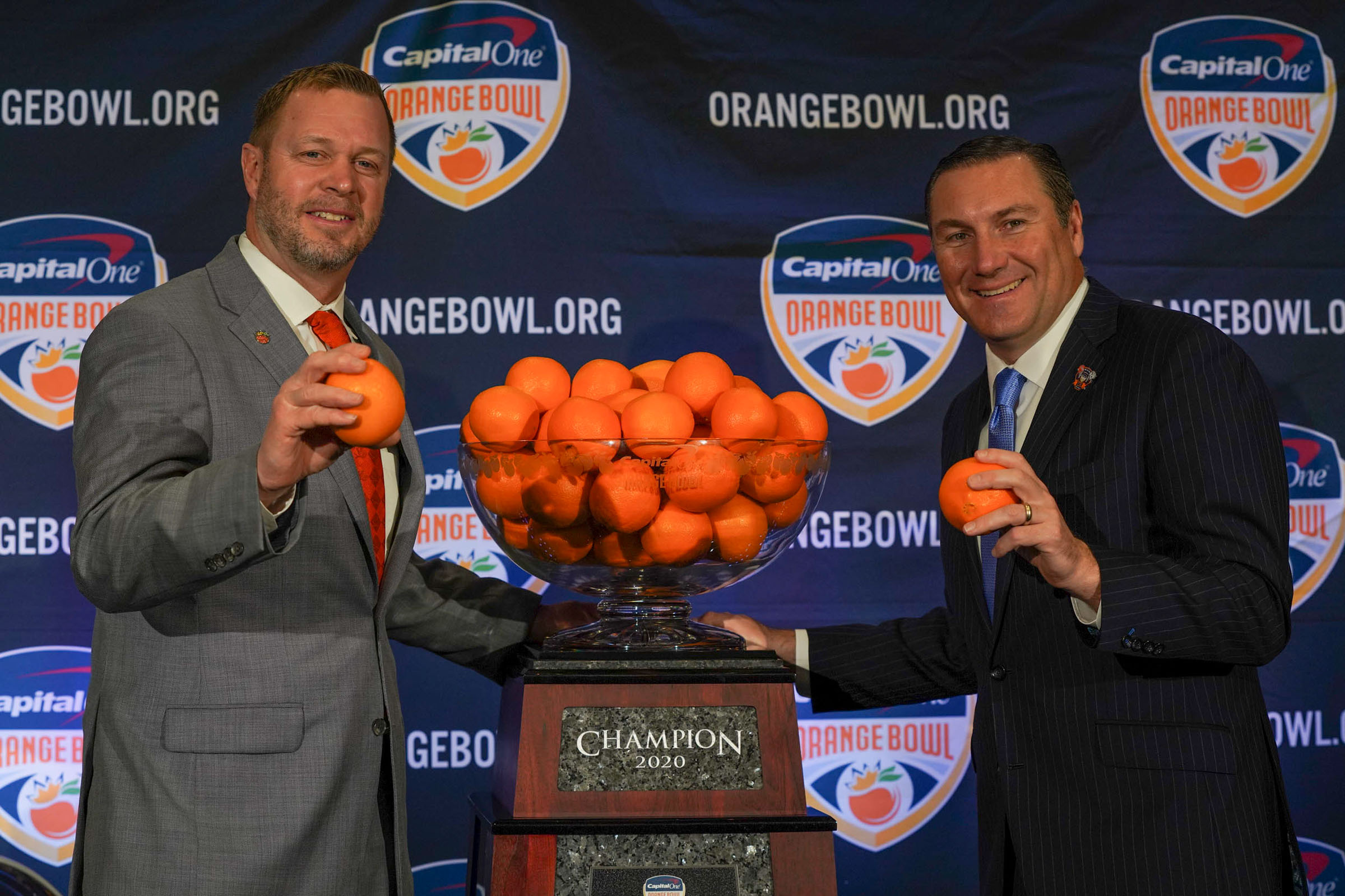Bronco Mendenhall, left, and Dan Mullen, right pose next to a glass bowl of oranges while each holding 1 orange
