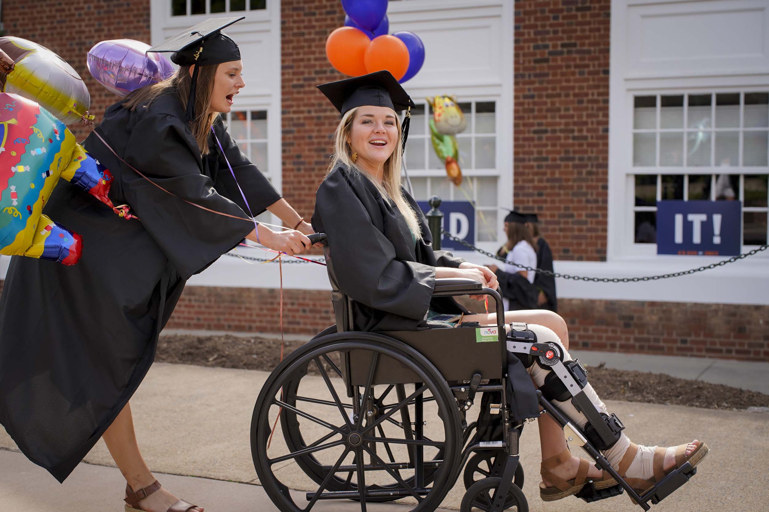 Graduate in a wheelchair being pushed by another graduate with balloons on the handles of the wheelchair