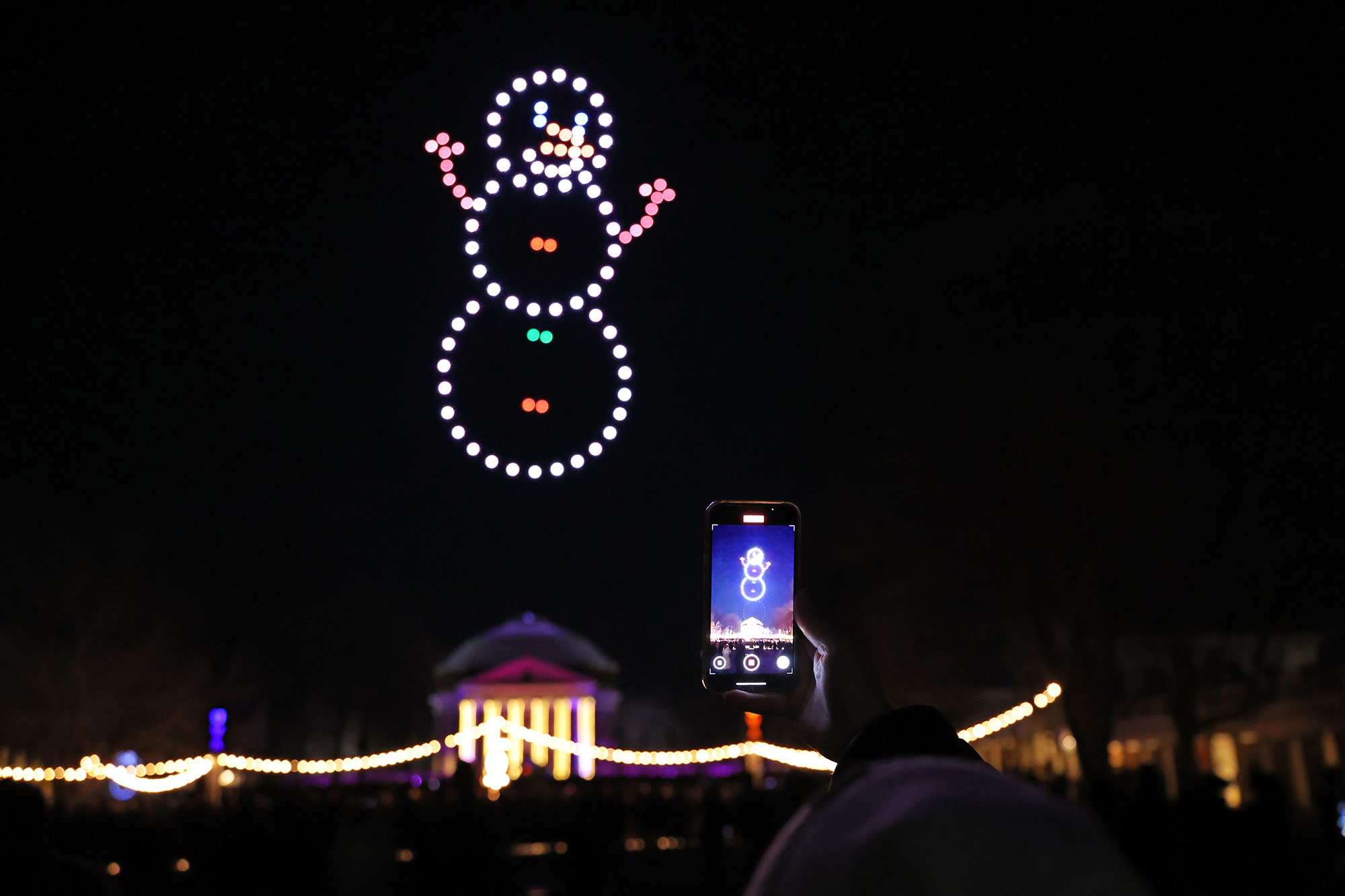 A person photographs a snowman made of lights on drones in the sky above the Rotunda.