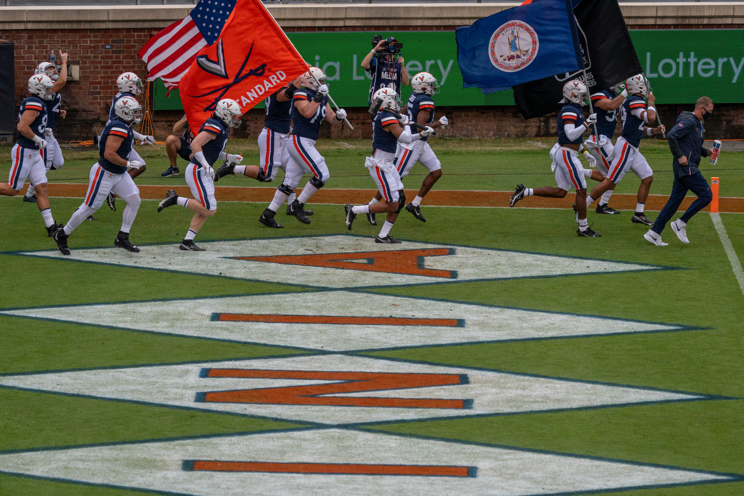 UVA Football team running on the field with the American flag and UVA flag