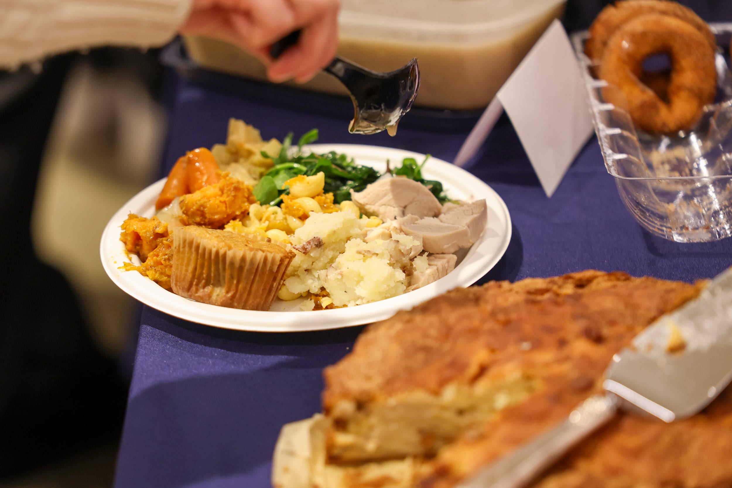 A close-up of a plate filled with various foods at a buffet-style meal, including turkey, mashed potatoes, vegetables, and a muffin. A hand holds a serving spoon above the plate, and additional dishes and desserts are visible on the table in the background.