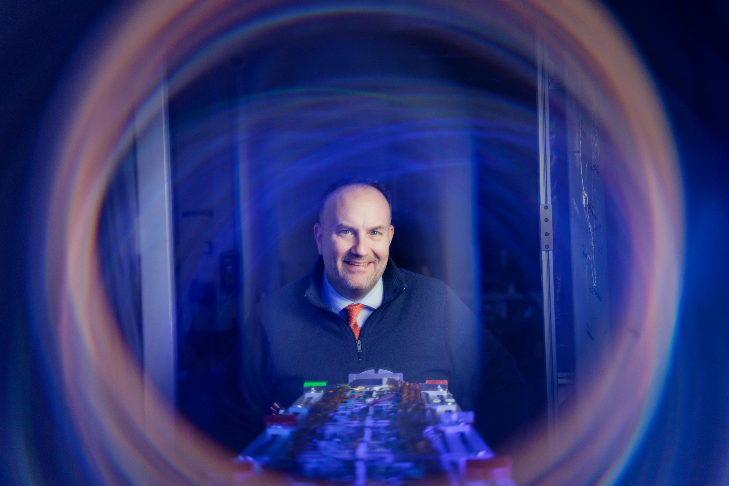 Portrait of Chris Neu behind technical equipment, framed by a circular colorful light effect.