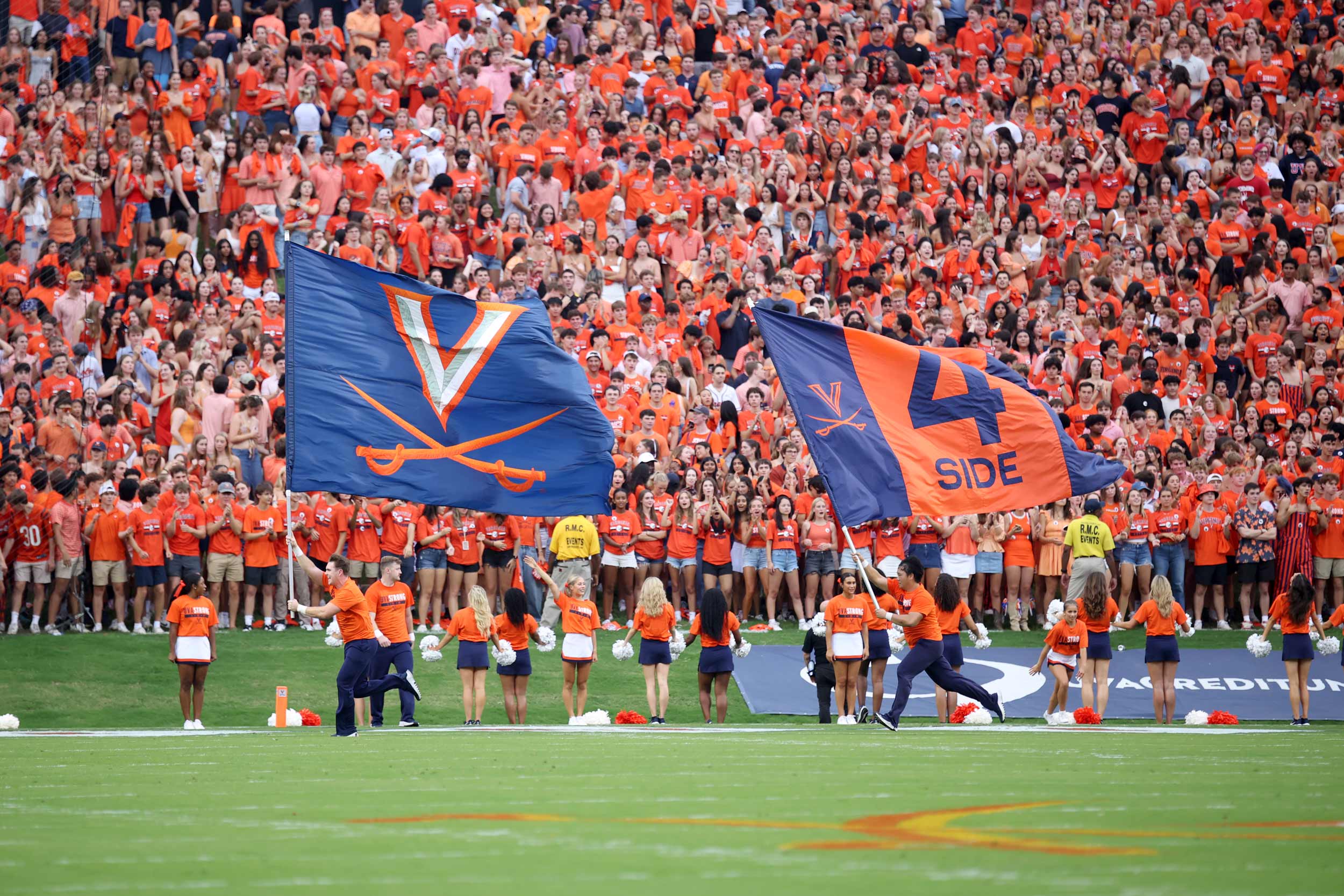 Football stadium full of fans wearing orange and waving two Virginia flags