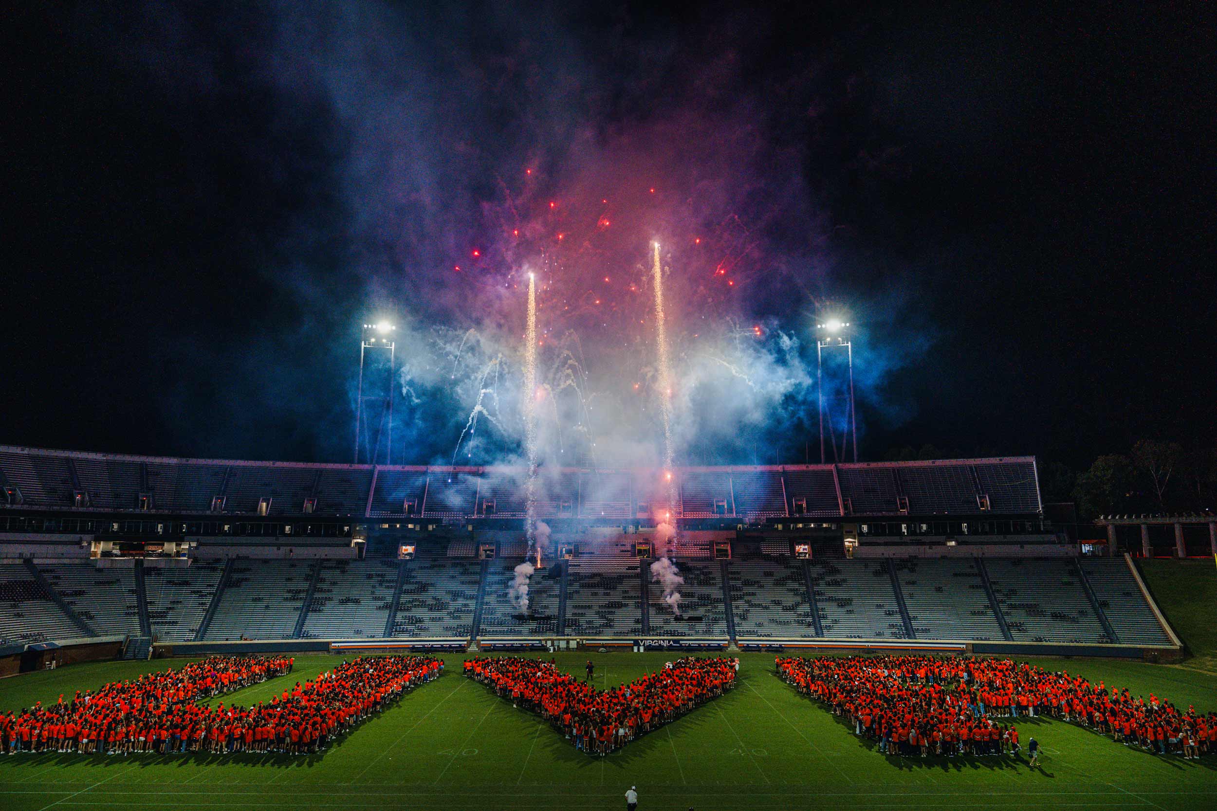 several people spell out the letters UVA on the field of Scott Stadium with fireworks in the background