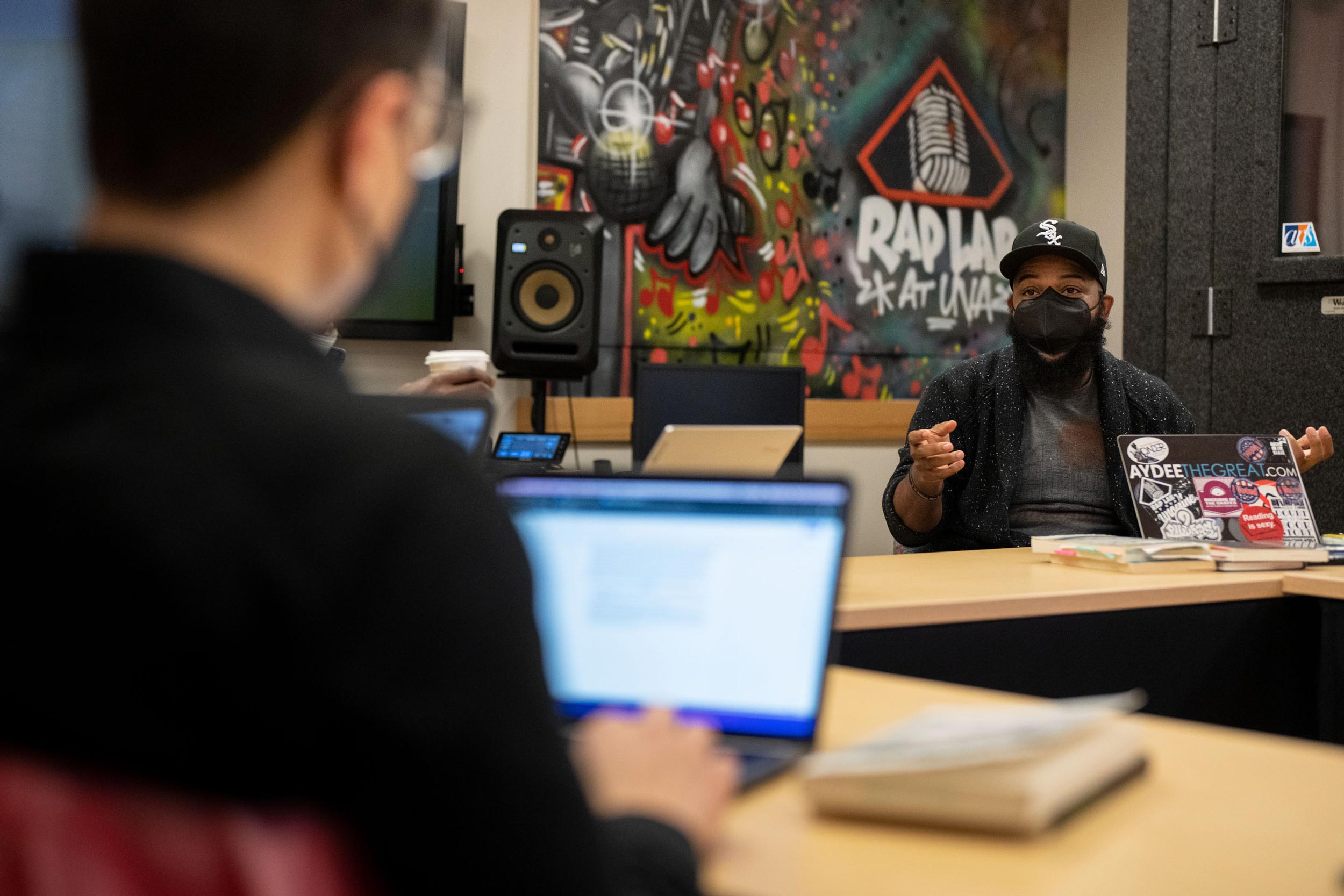 A man sits at a long table in front of a graffiti-style mural reading Rap Lab. He speaks to a student with a laptop.
