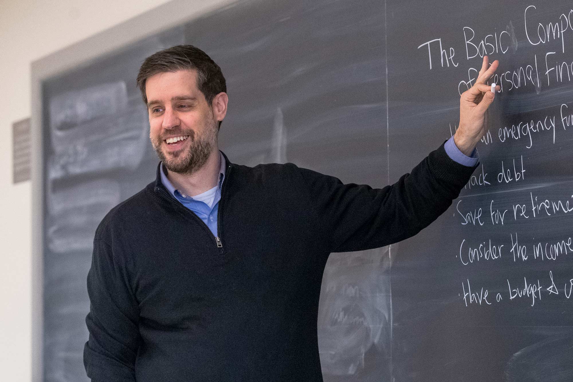 UVA associate professor Andrew Pennock, smiling writes on a blackboard: The basic components of personal finance.