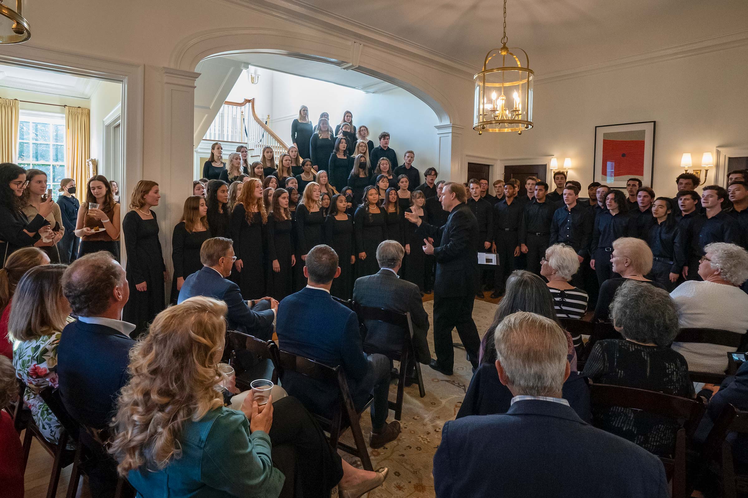 A large choir dressed in black fills a stairwell and back of a room singing for a small seated audience