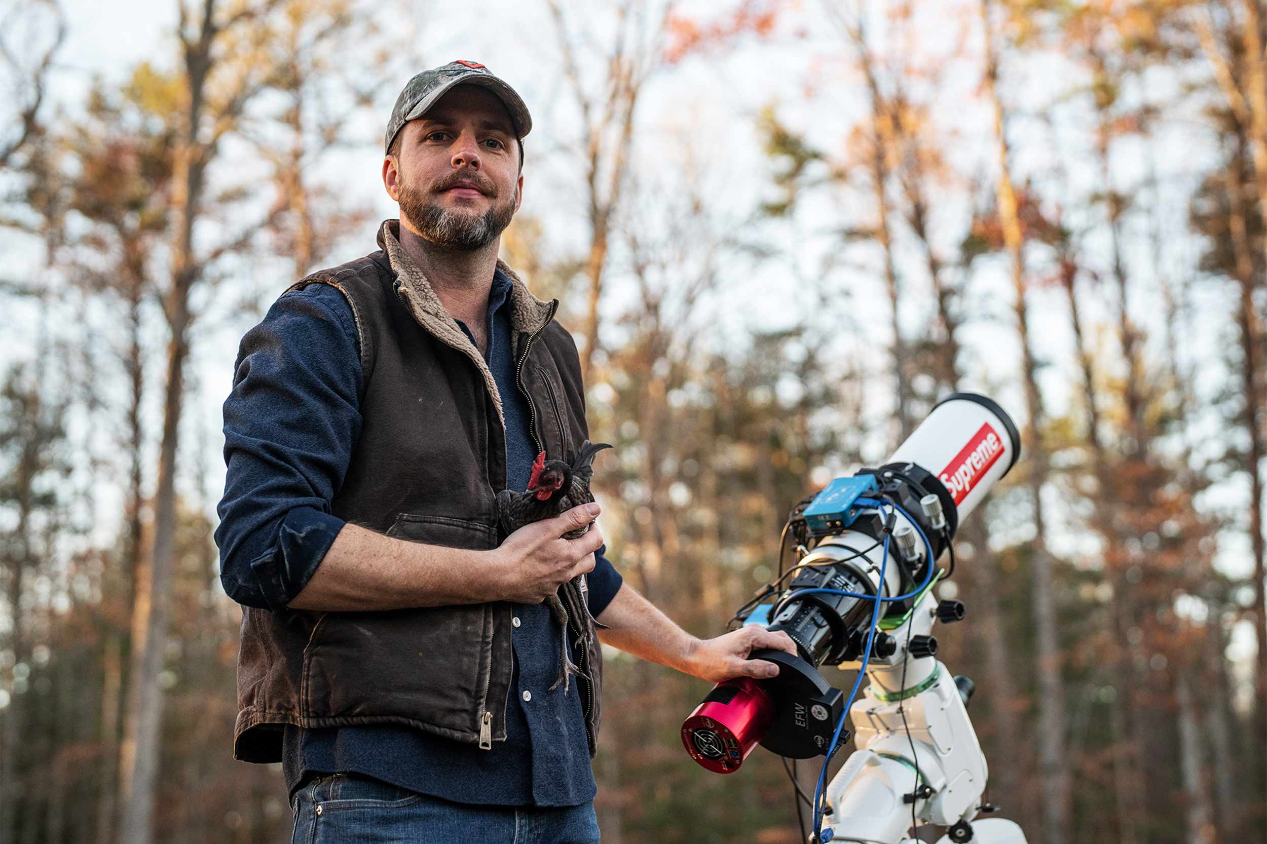 Brennan Gilmore stands in the woods with one hand on a telescope, the other holding a small black chicken