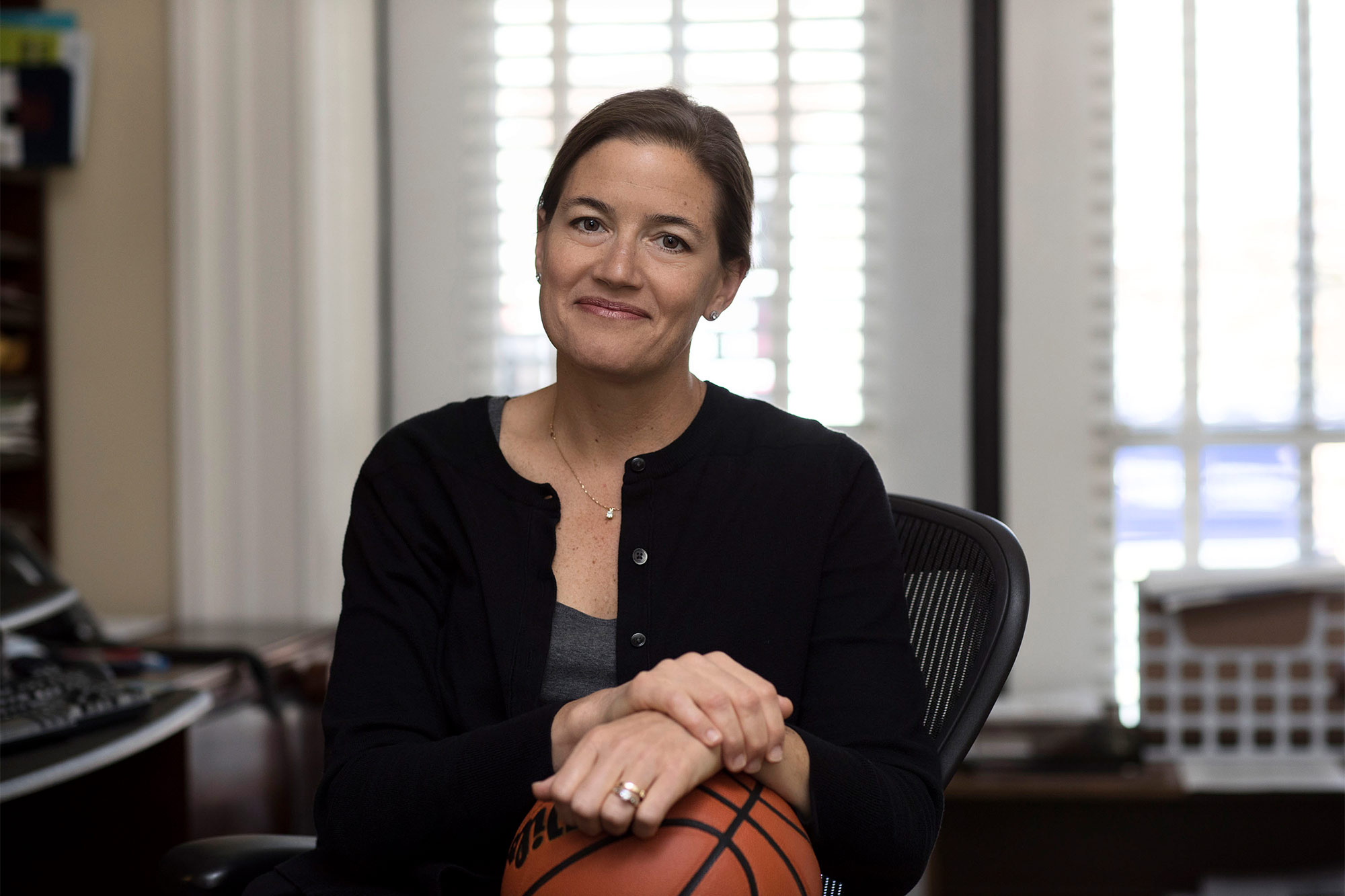 Carrie Heilman sits at a desk, with her hands resting on a basketball in her lap, and smiles at the camera