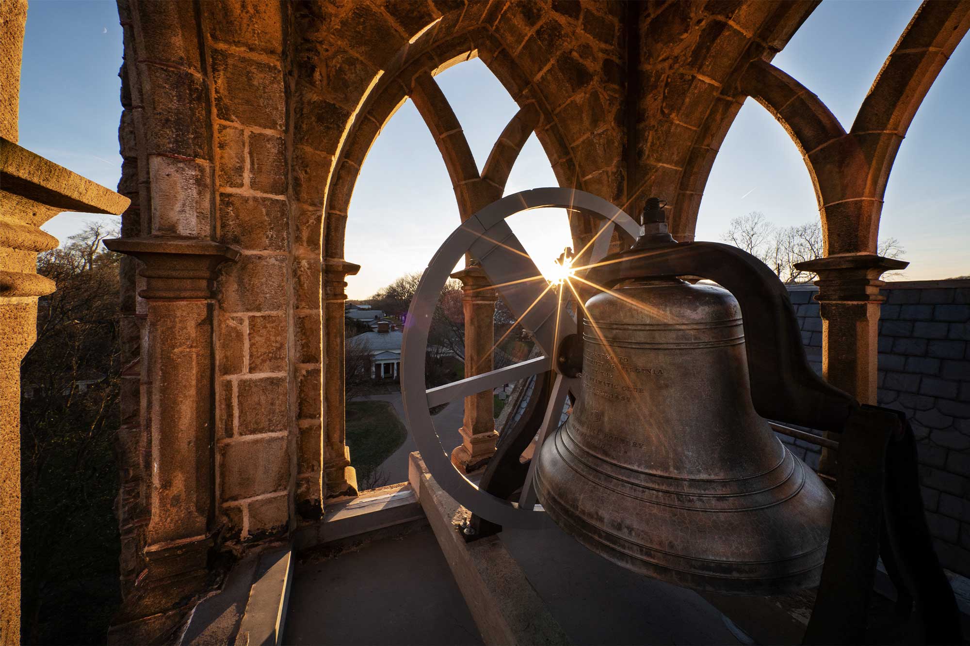 The sun shines from behind the large bell in the tower of the UVA chapel.