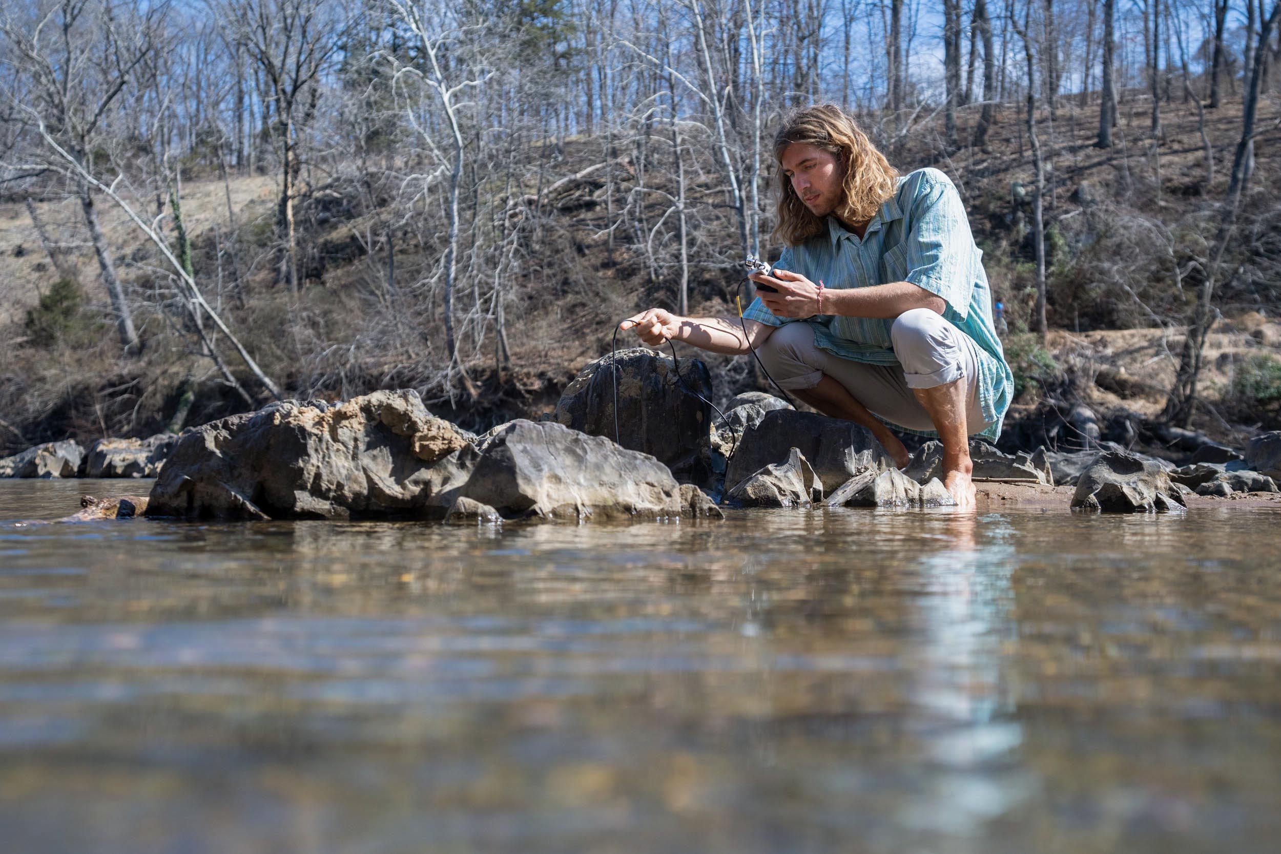 UVA student Davis Coffee squats next to a creek, holding a device whose cable runs into the water
