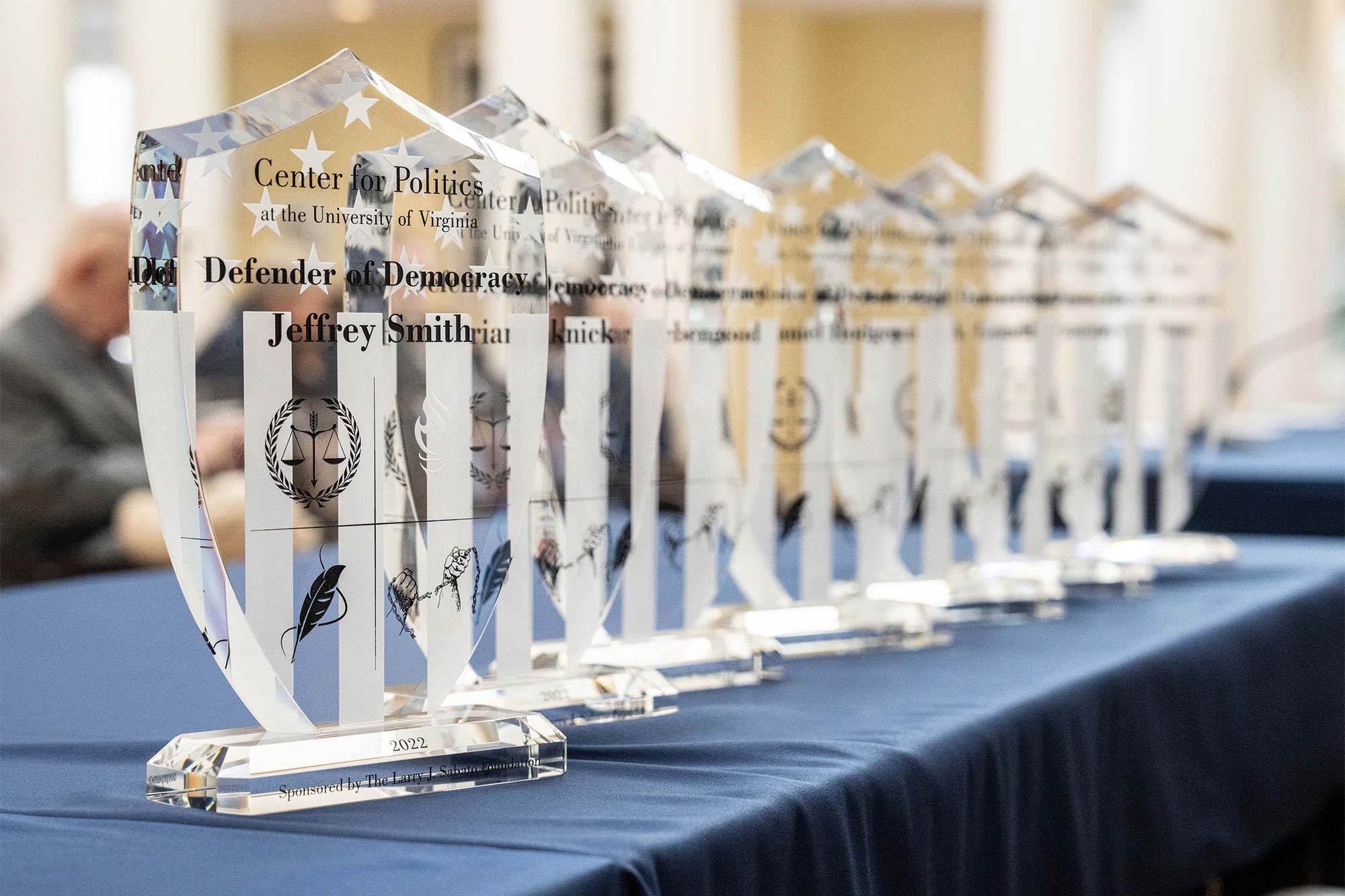Seven glass awards lined up on a table with a blue tablecloth