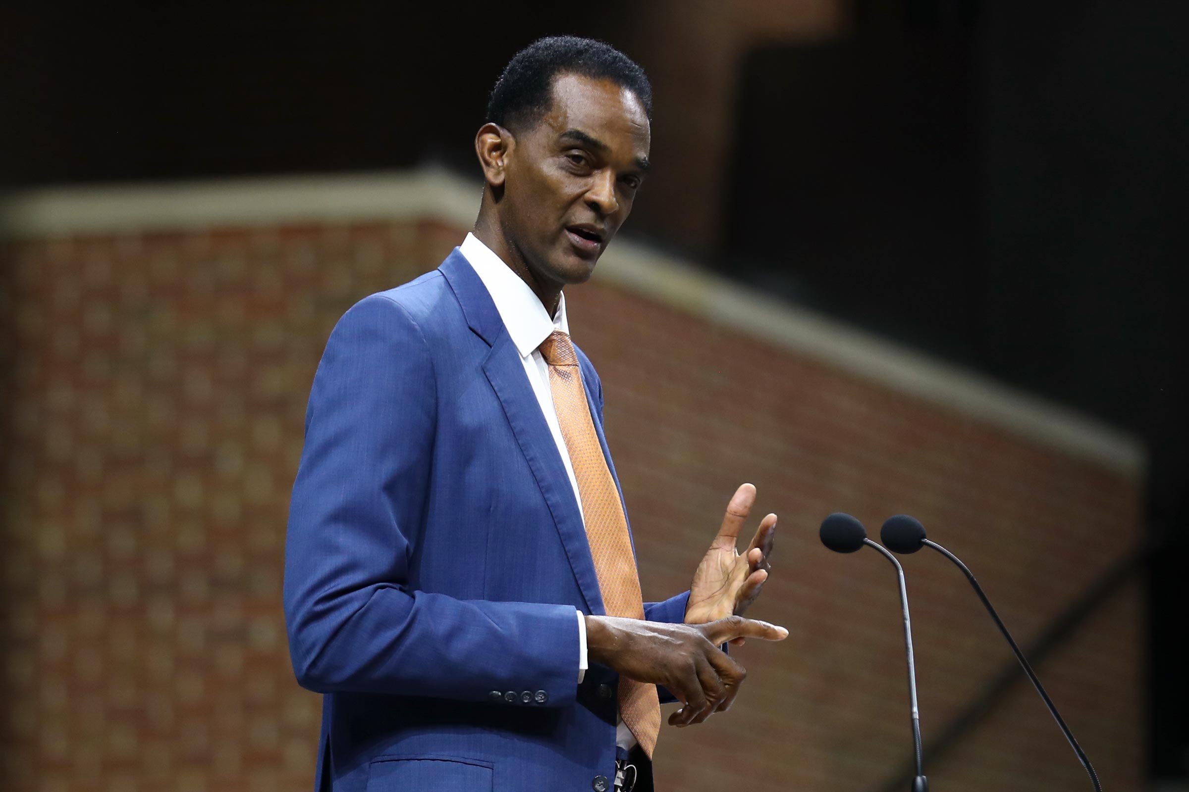 Ralph Sampson, in a blue suit and orange tie, gives a speech at a podium