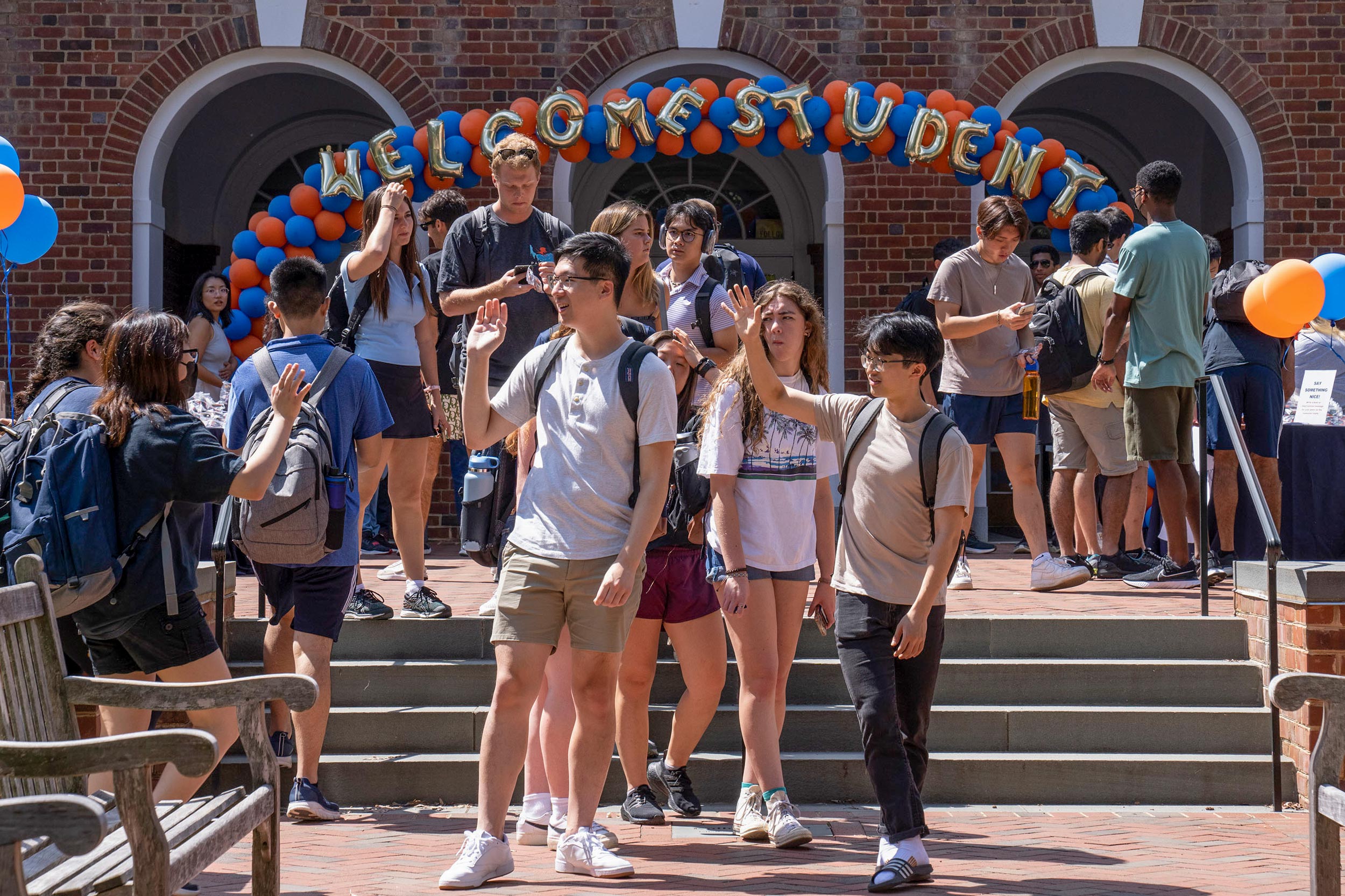 Students pass each other and wave under an orange and blue ballon arch with silver balloon letters spelling out Welcome Students