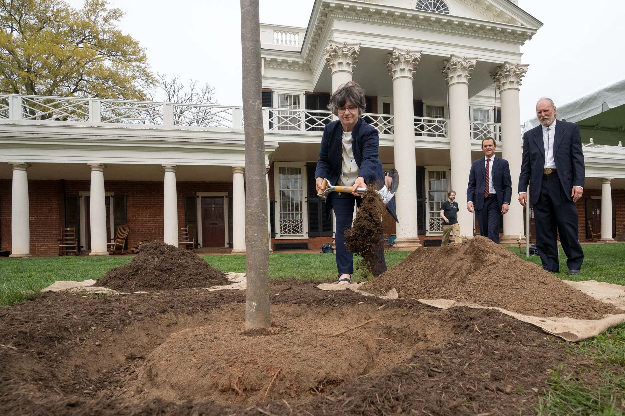 Mary Hughes shovels a pile of soil into a planting hole. Worthy Martin and UVA President Jim Ryan look on.