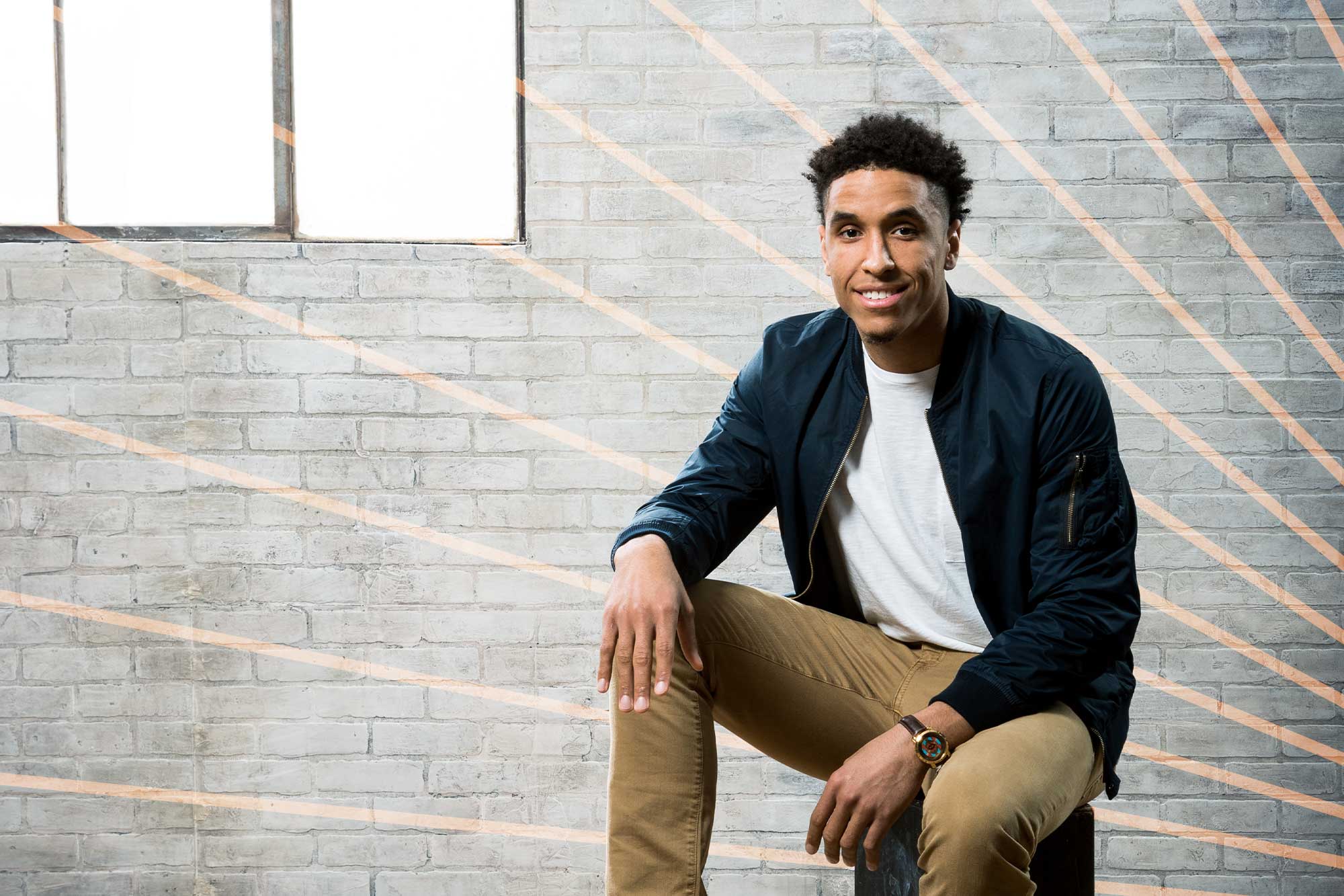Malcolm Brogdon sits casually in front of a white brick wall and smiles at the camera. Orange lines radiate behind him.