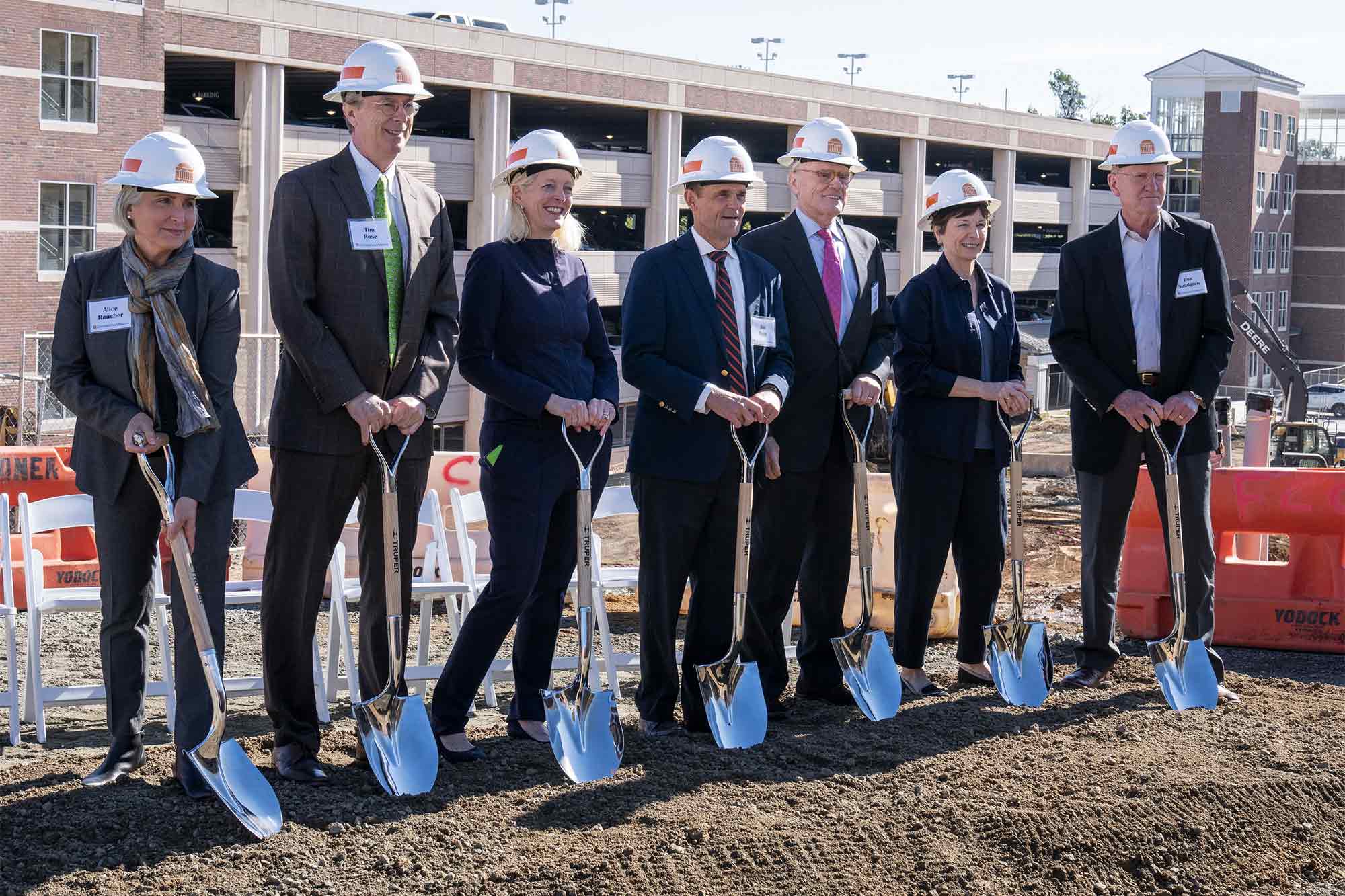 Seven adults in hard hats stand in a row, holding shiny shovels and posing for a group shot