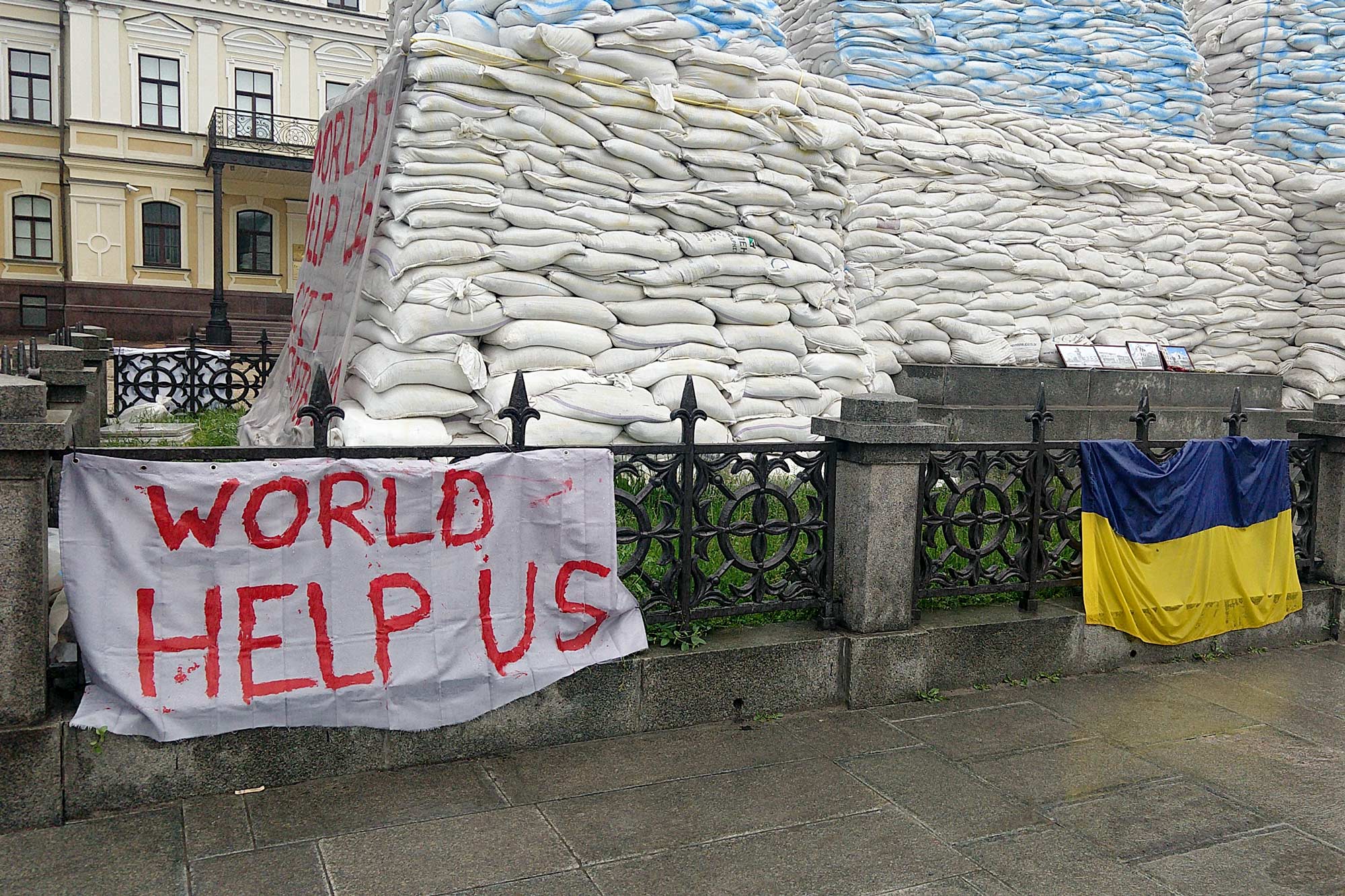 A white sheet with 'World Help Us' painted in red letters hangs next to the flag of Ukraine in front of an structure of hundreds of sandbags