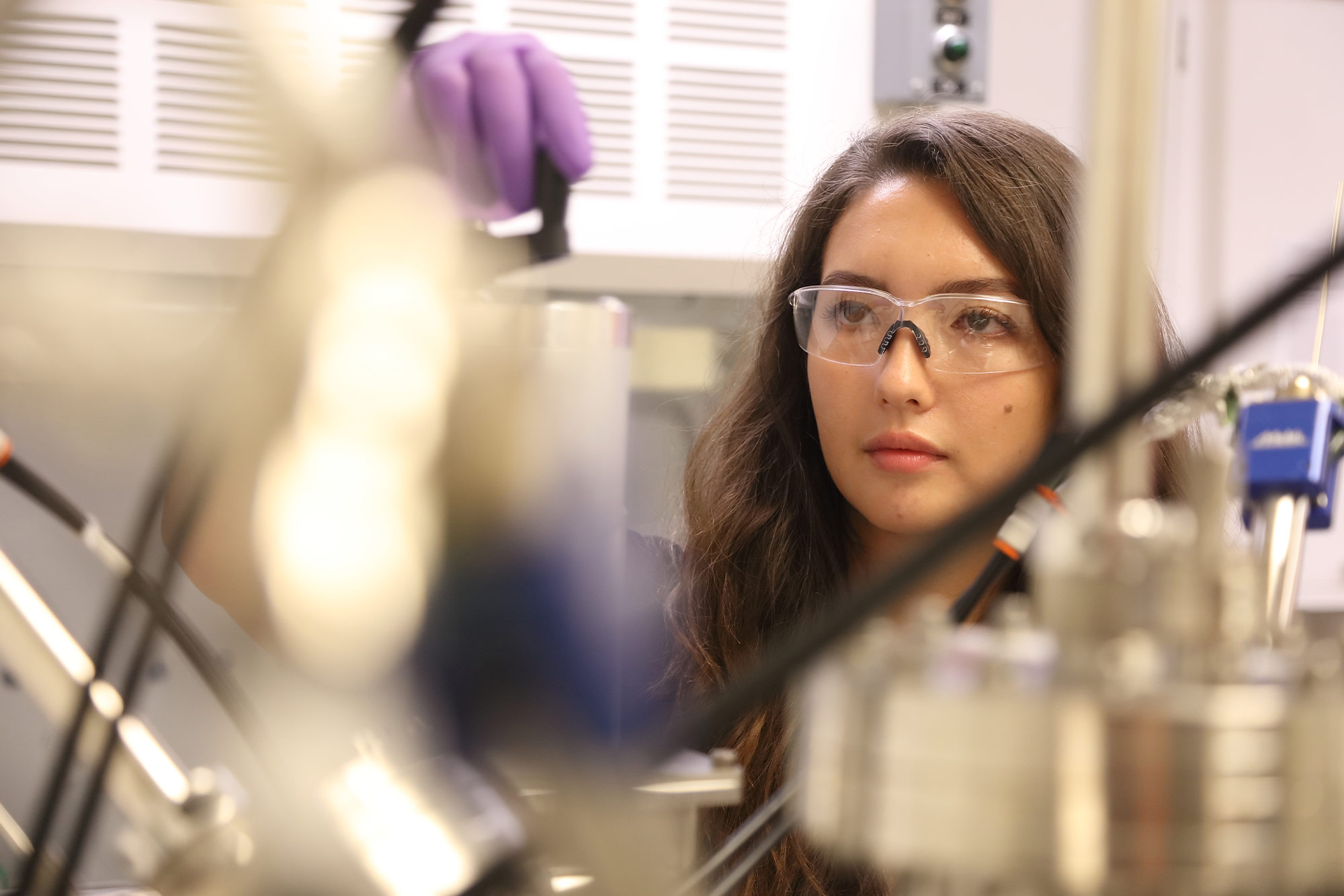 A woman in safety goggles and purple latex gloves adjusts a piece of lab equipment